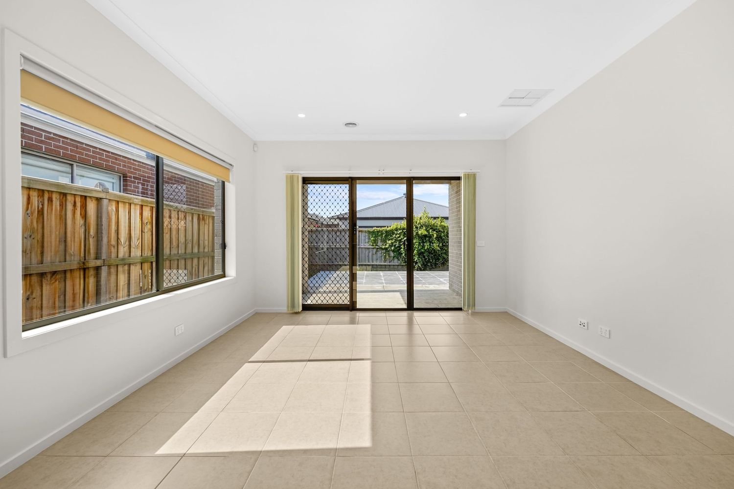 Empty room with beige tile floors, large windows, and sliding glass doors opening to a patio with wooden fence views.