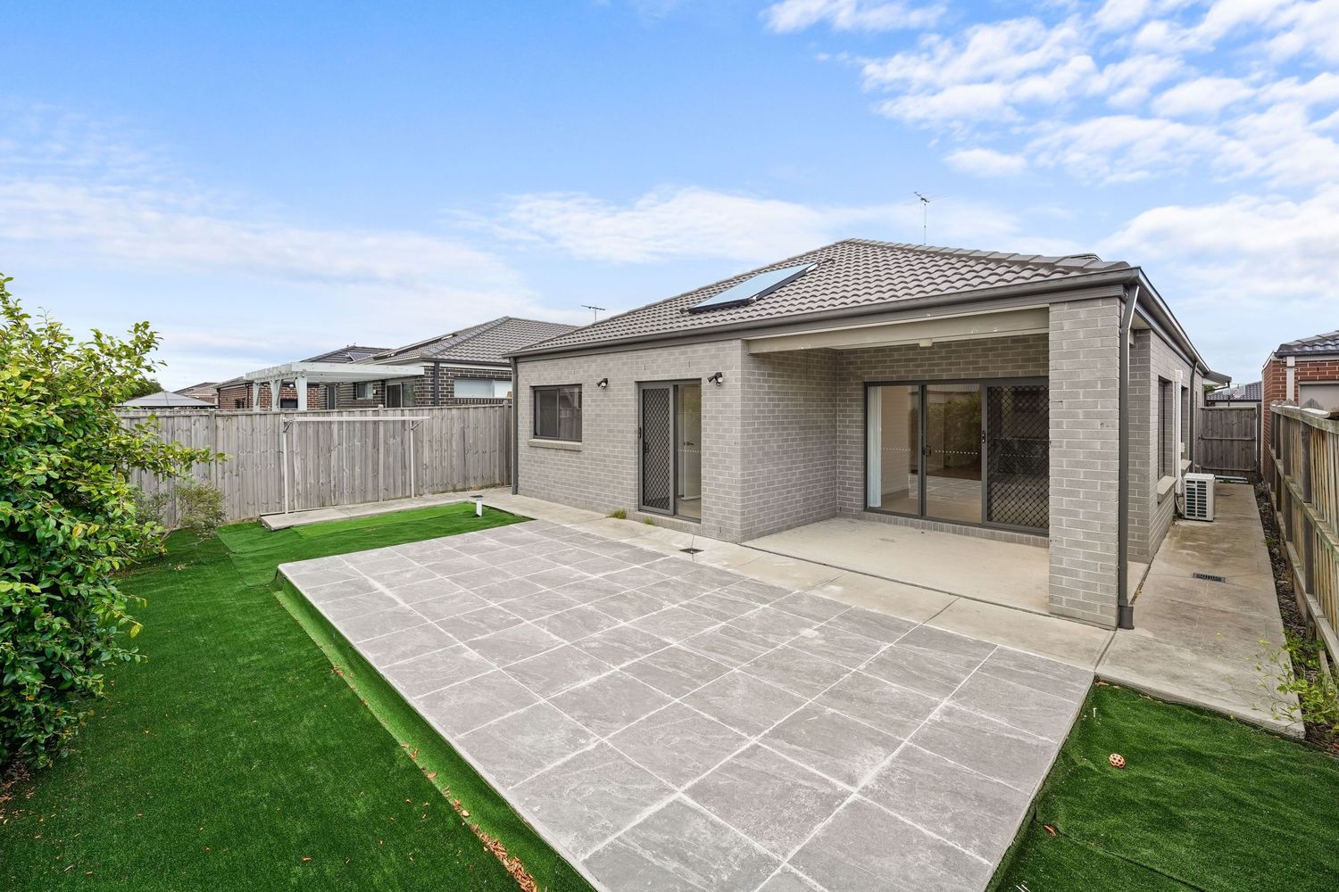 Modern single story house with gray exterior and large paved patio area overlooking well-maintained lawn under blue sky.