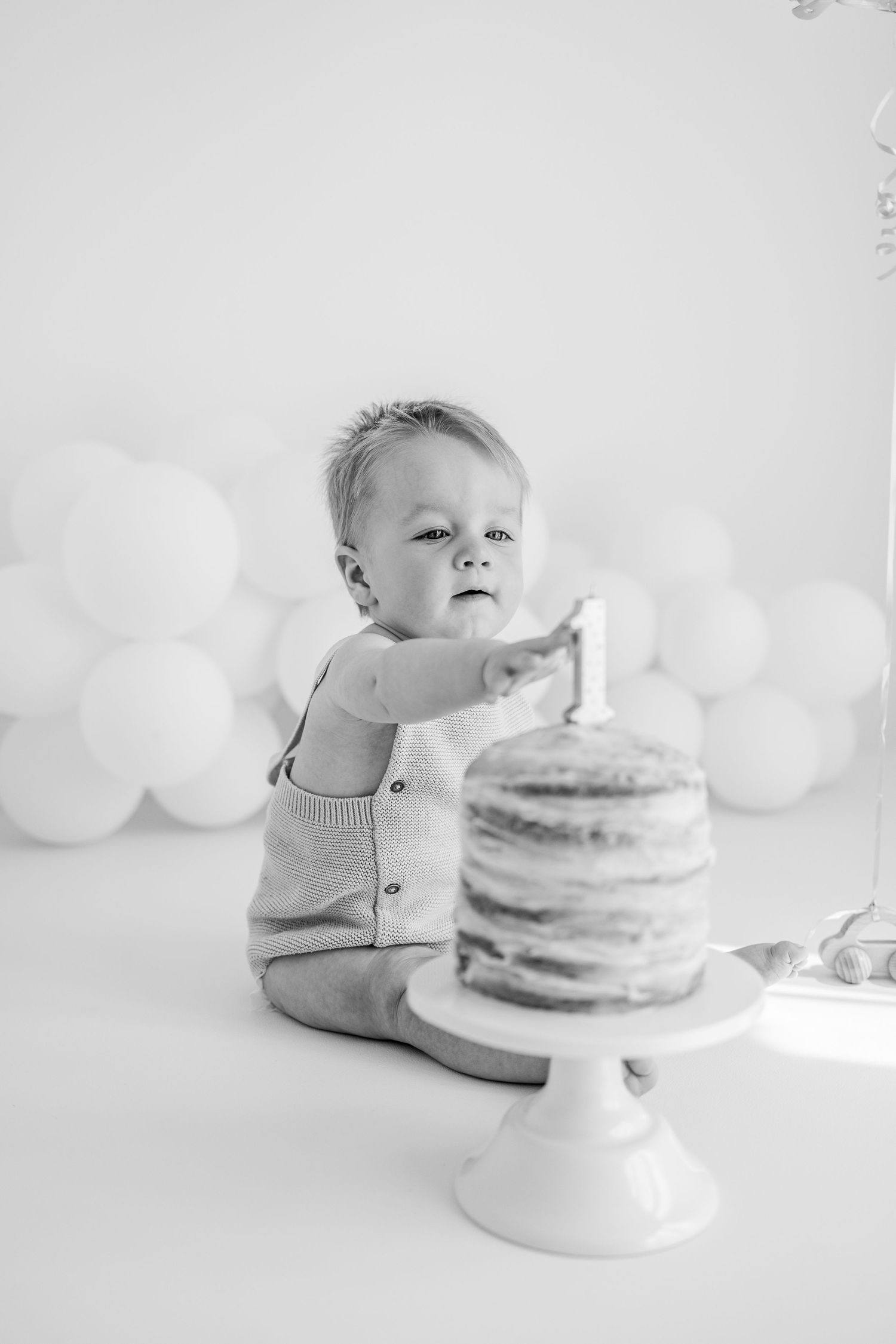 A small child sits in a large white basin surrounded by sage green and cream colored balloons in a soft studio setting.