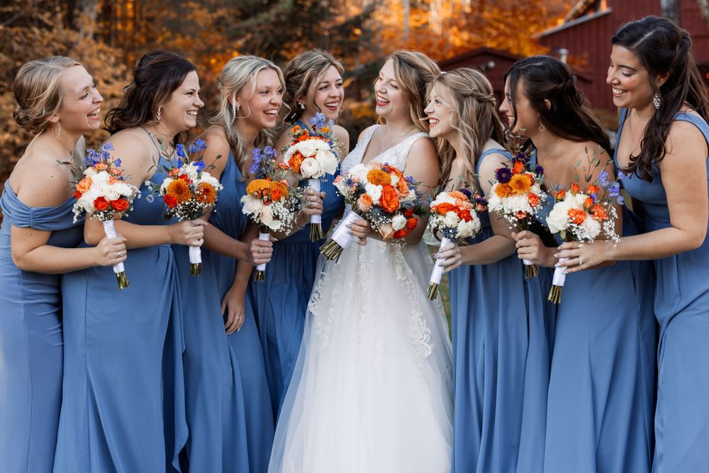 Bridesmaids in blue dresses stand with bride holding orange and white floral bouquets.