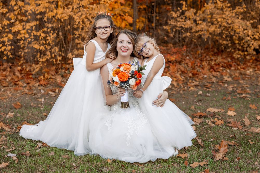 Three figures in white dresses pose together on autumn leaves holding orange bouquets.