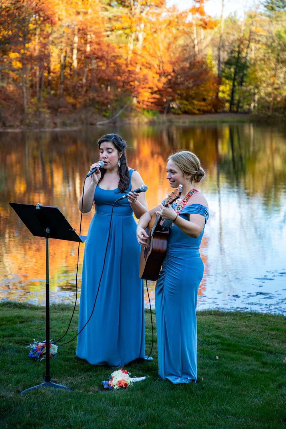 Musicians in blue dresses perform by lakeside during autumn wedding ceremony.