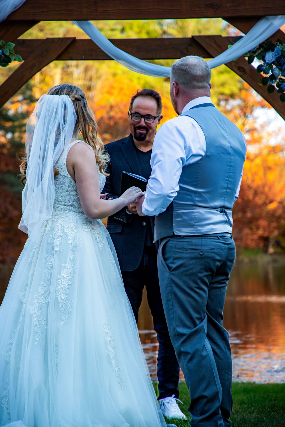 Wedding ceremony under wooden arch with officiant and couple by autumn lake.