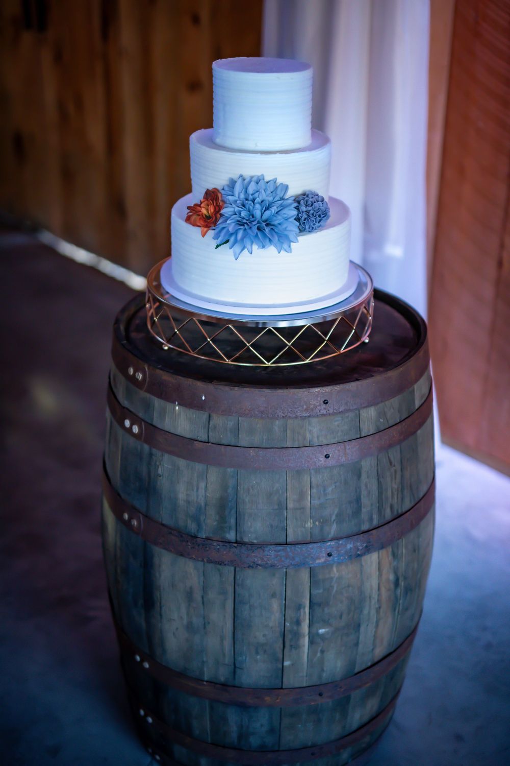 Three-tiered white wedding cake with blue and red flowers displayed on a rustic wooden barrel.