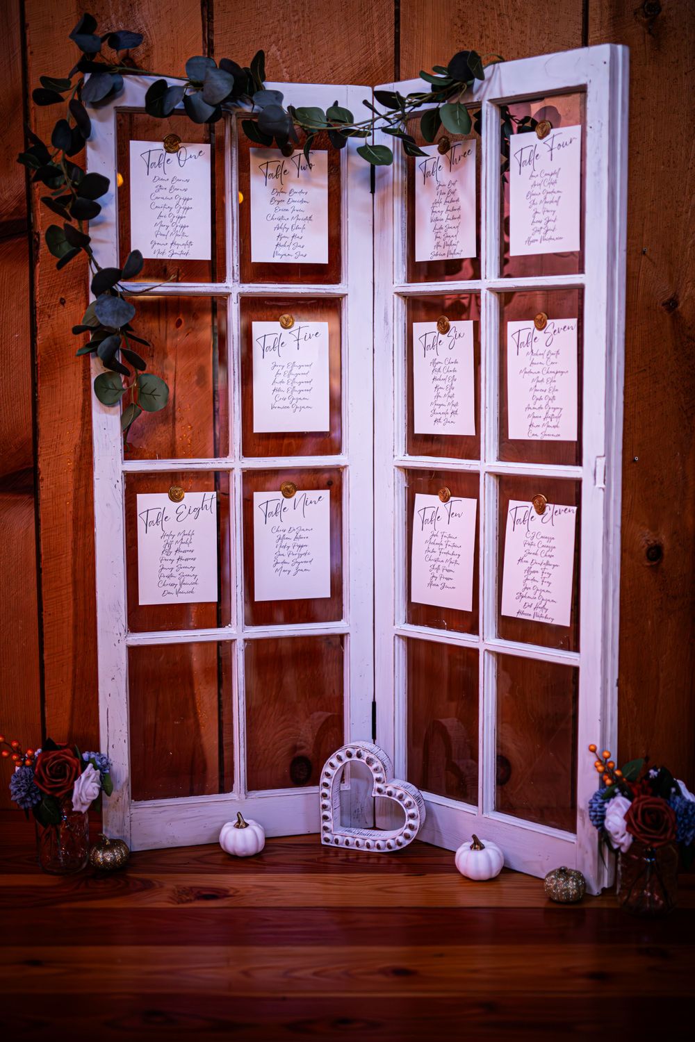 Wedding seating chart displayed on white window panes decorated with greenery and small pumpkins.