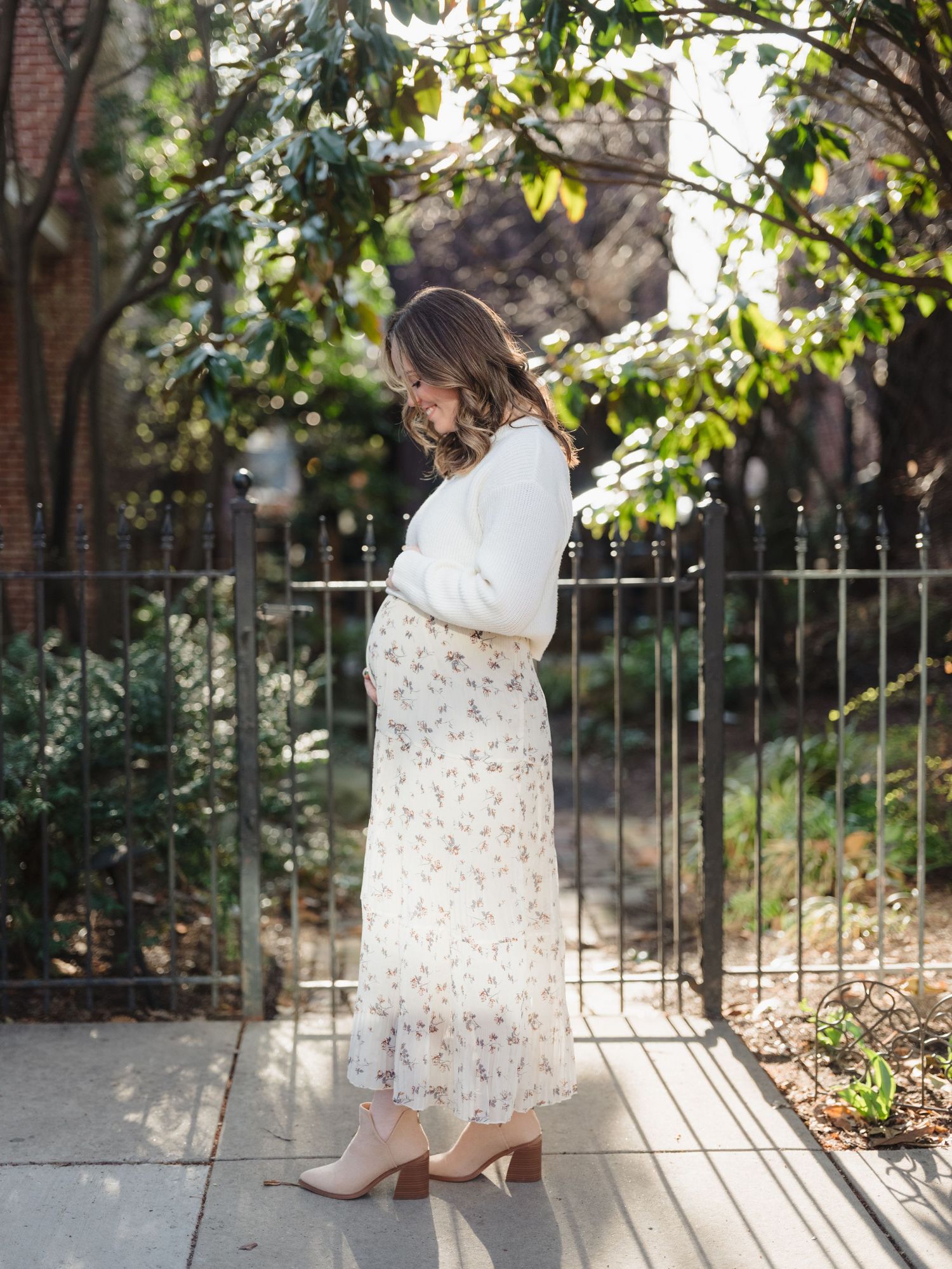 A person in a white sweater and floral skirt stands beside an iron gate in a sunlit garden setting.