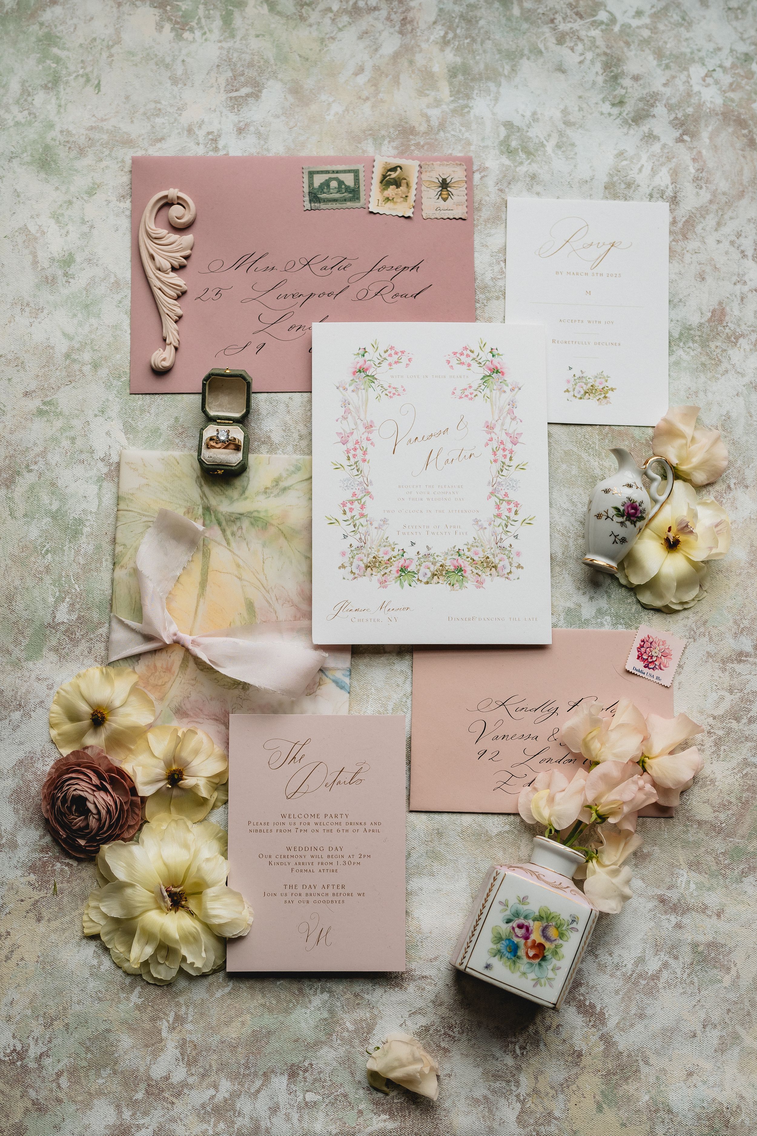 Flat lay of vintage-style wedding invitation suite in blush pink with floral details, stamps, and scattered cream flowers.