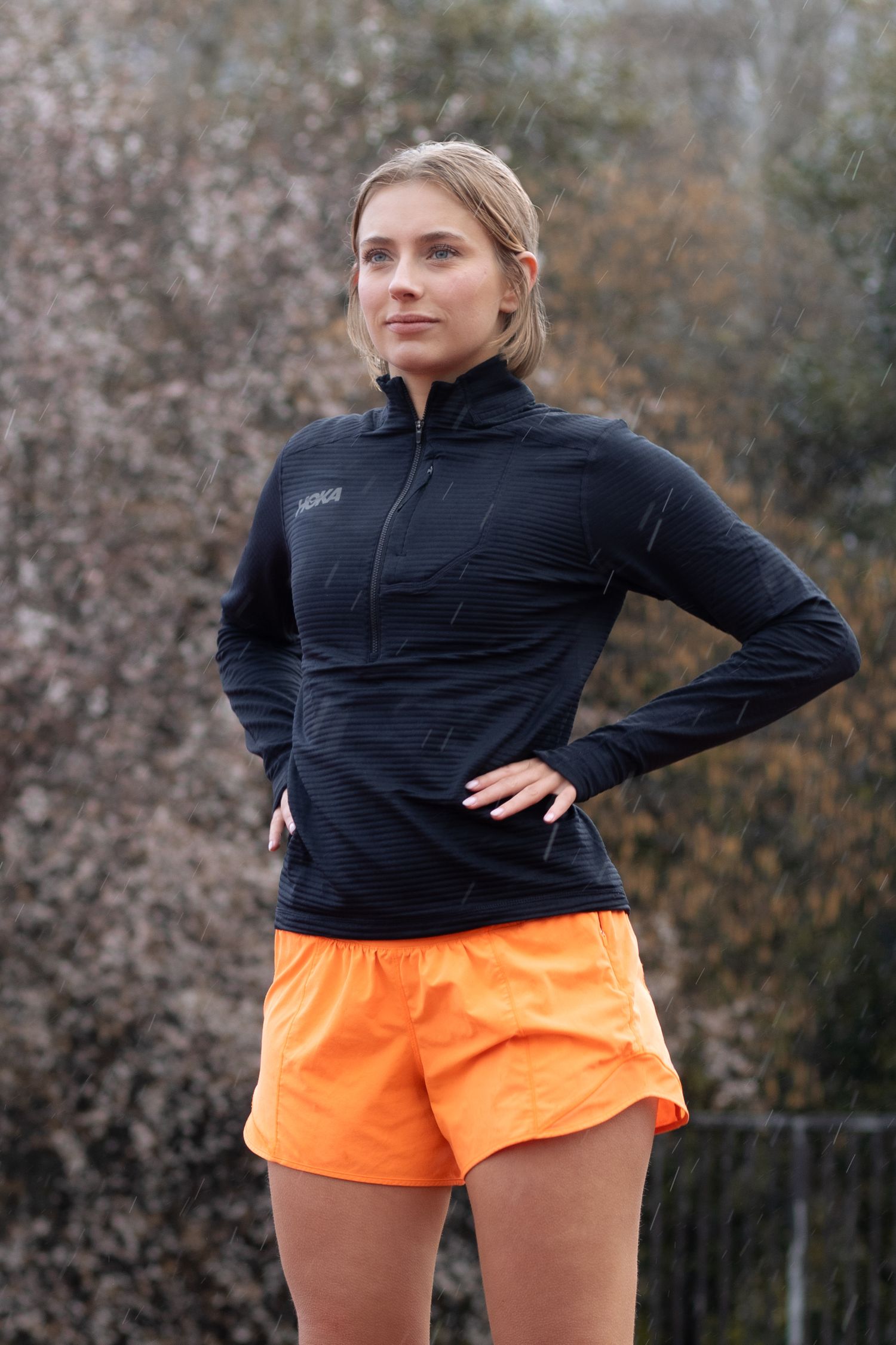 Athletic person in black jacket and orange shorts posing outdoors.