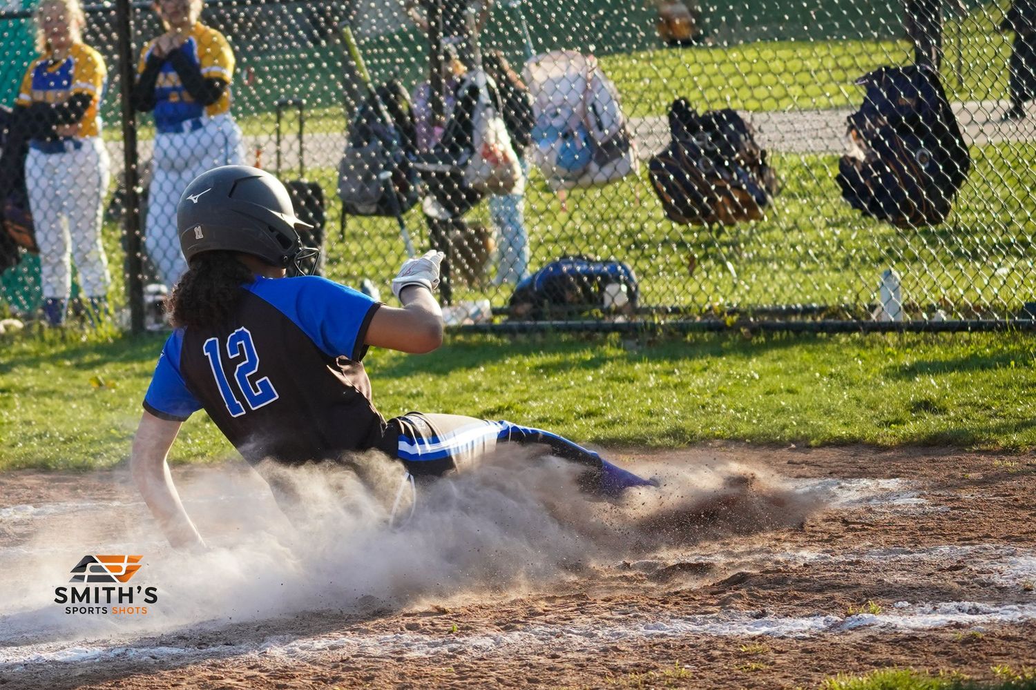 Baseball player in blue jersey number 12 slides dramatically into home plate creating a cloud of dust during game.