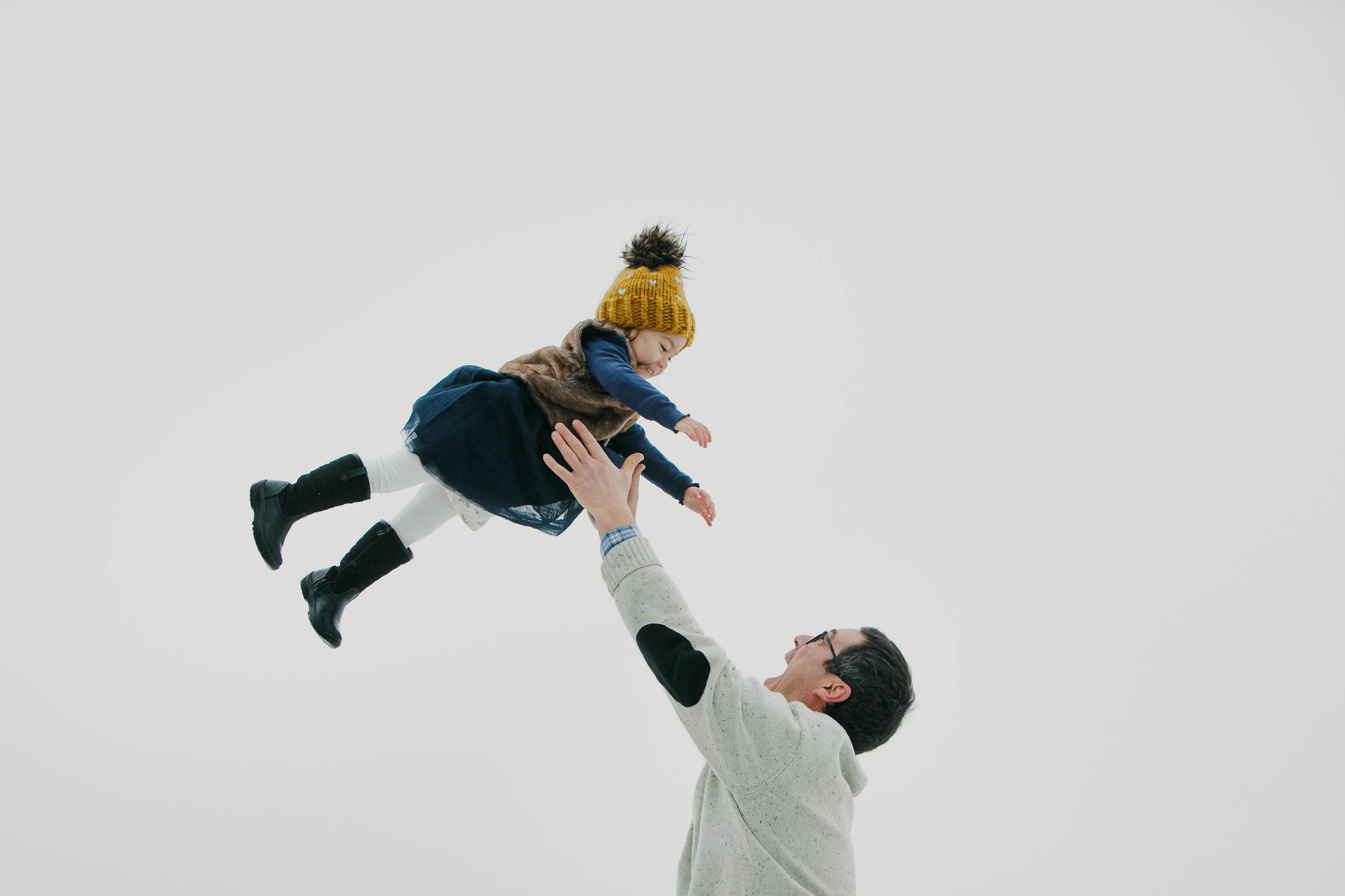 Person in white sweater lifts child wearing yellow beanie and blue coat against white background.