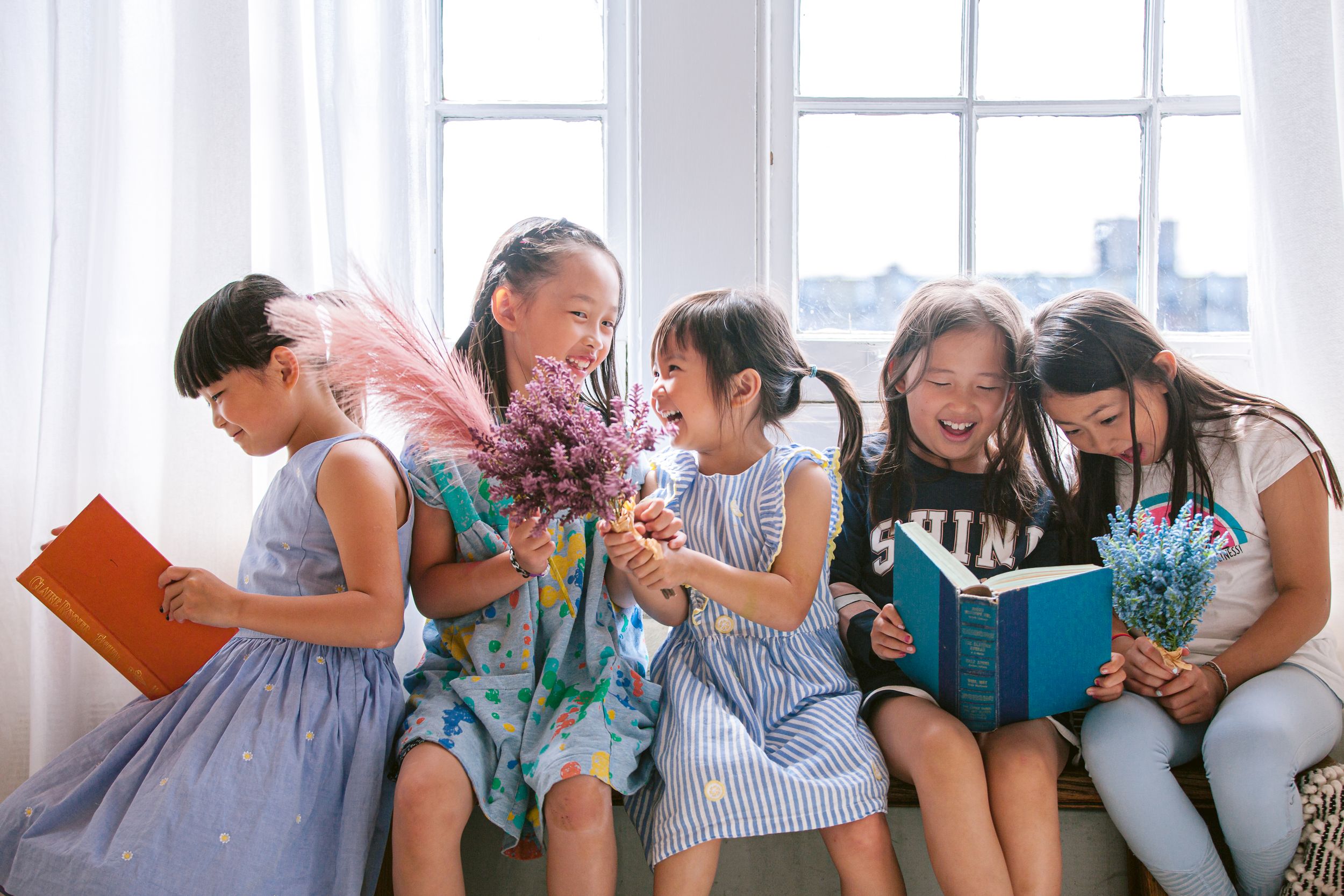 A group of friends share joyful laughter while reading books together on a bright window seat.