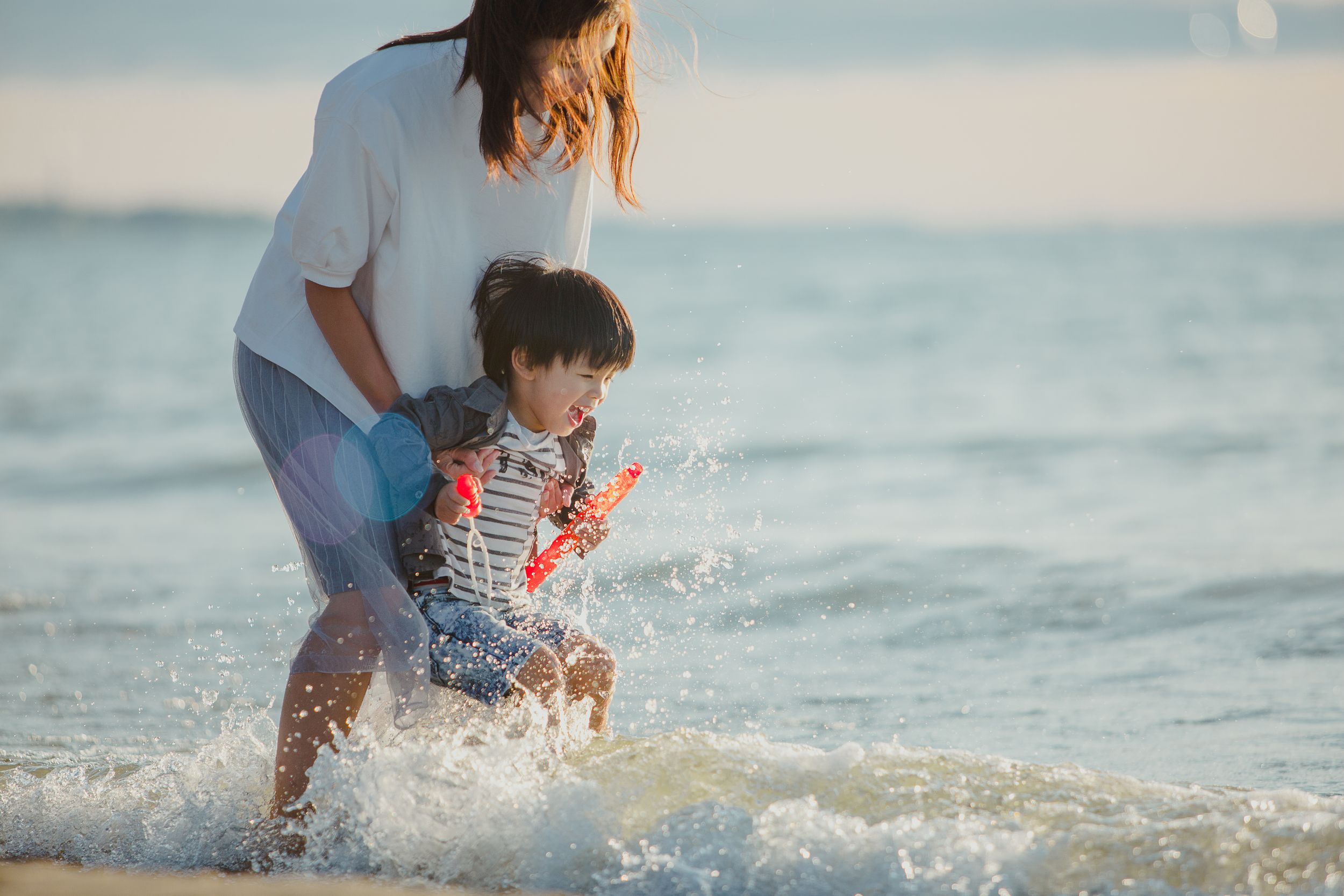 Parent and child splash and play together in gentle ocean waves at sunset wearing casual beach attire.