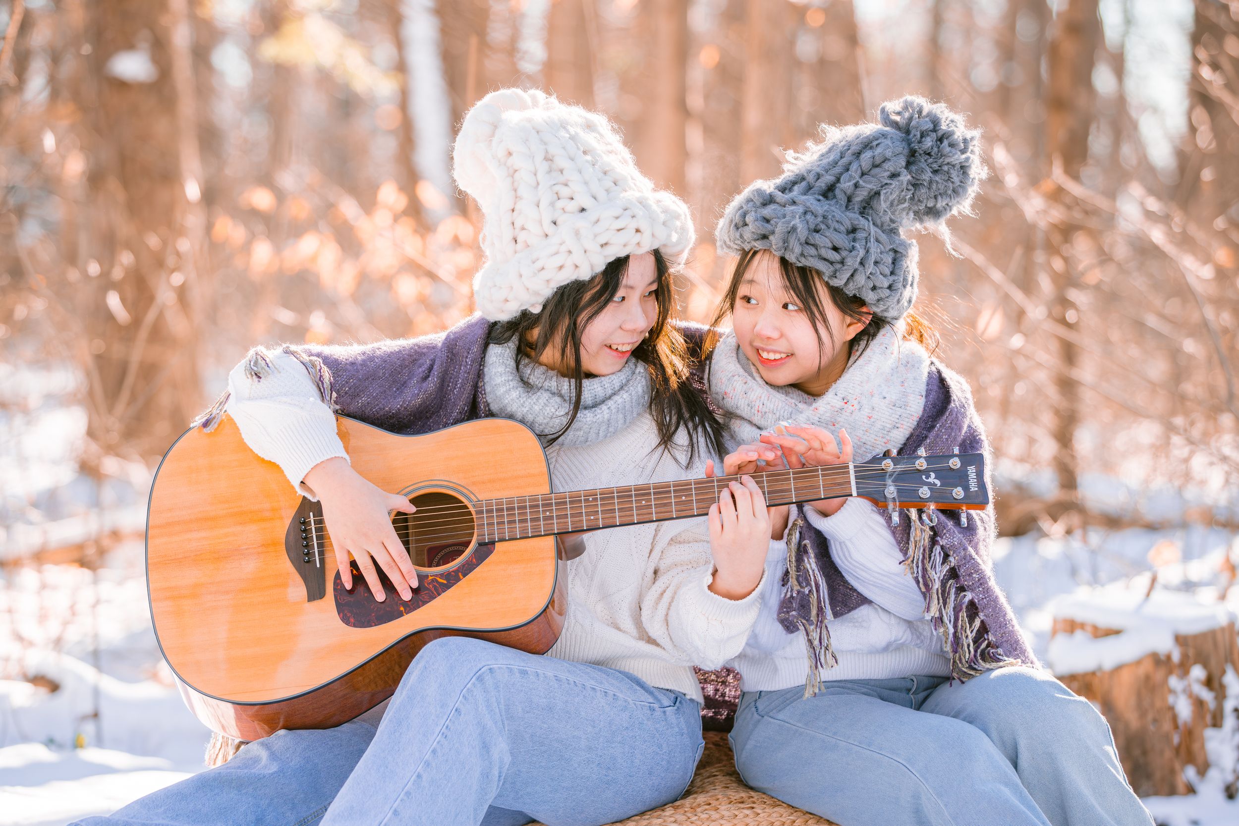 Two people sharing an acoustic guitar while sitting together outdoors in a snowy winter setting wearing knit hats.