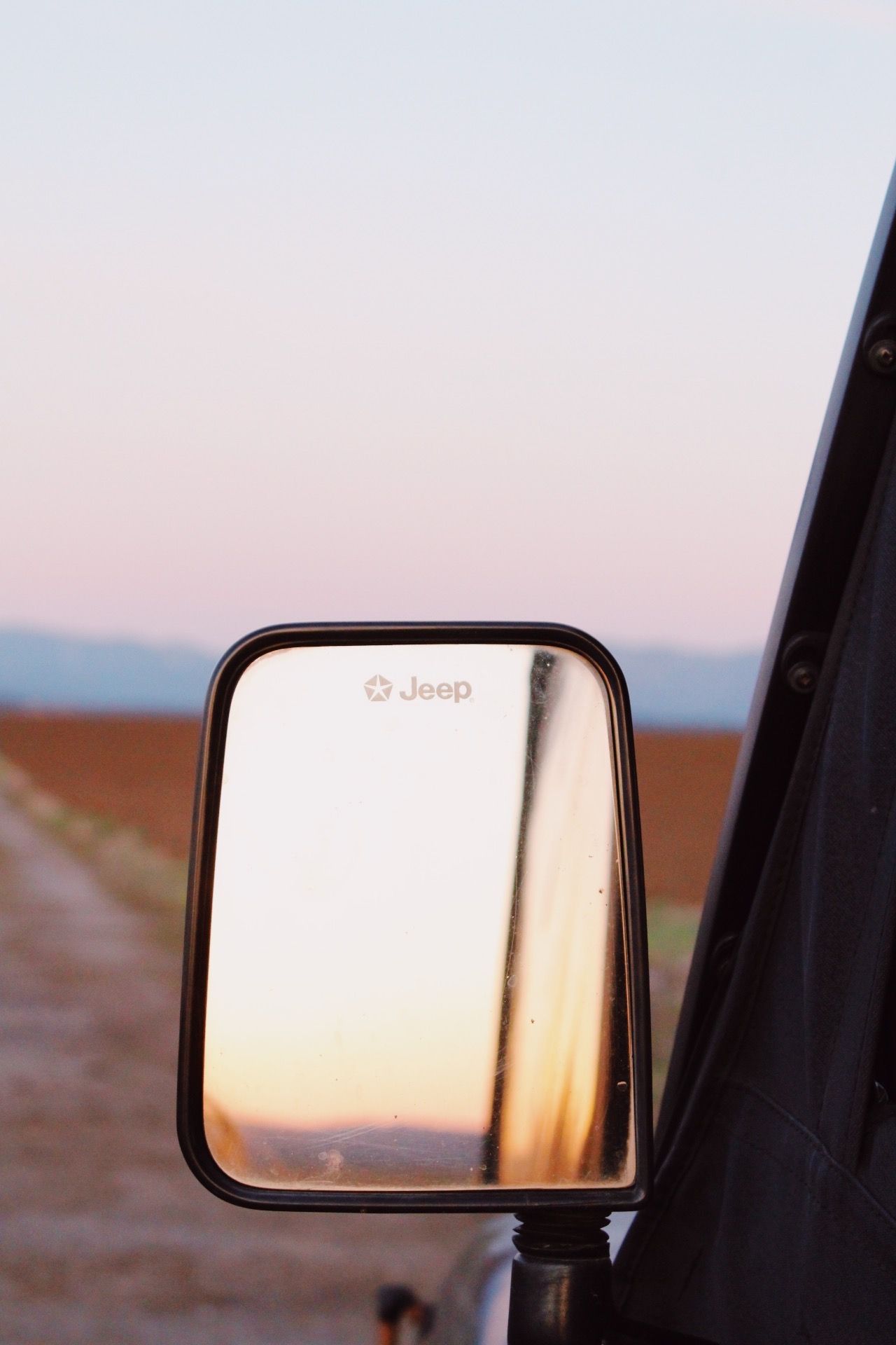 Sunset reflected in a Jeep side mirror on a rural dirt road against a pink and blue sky.