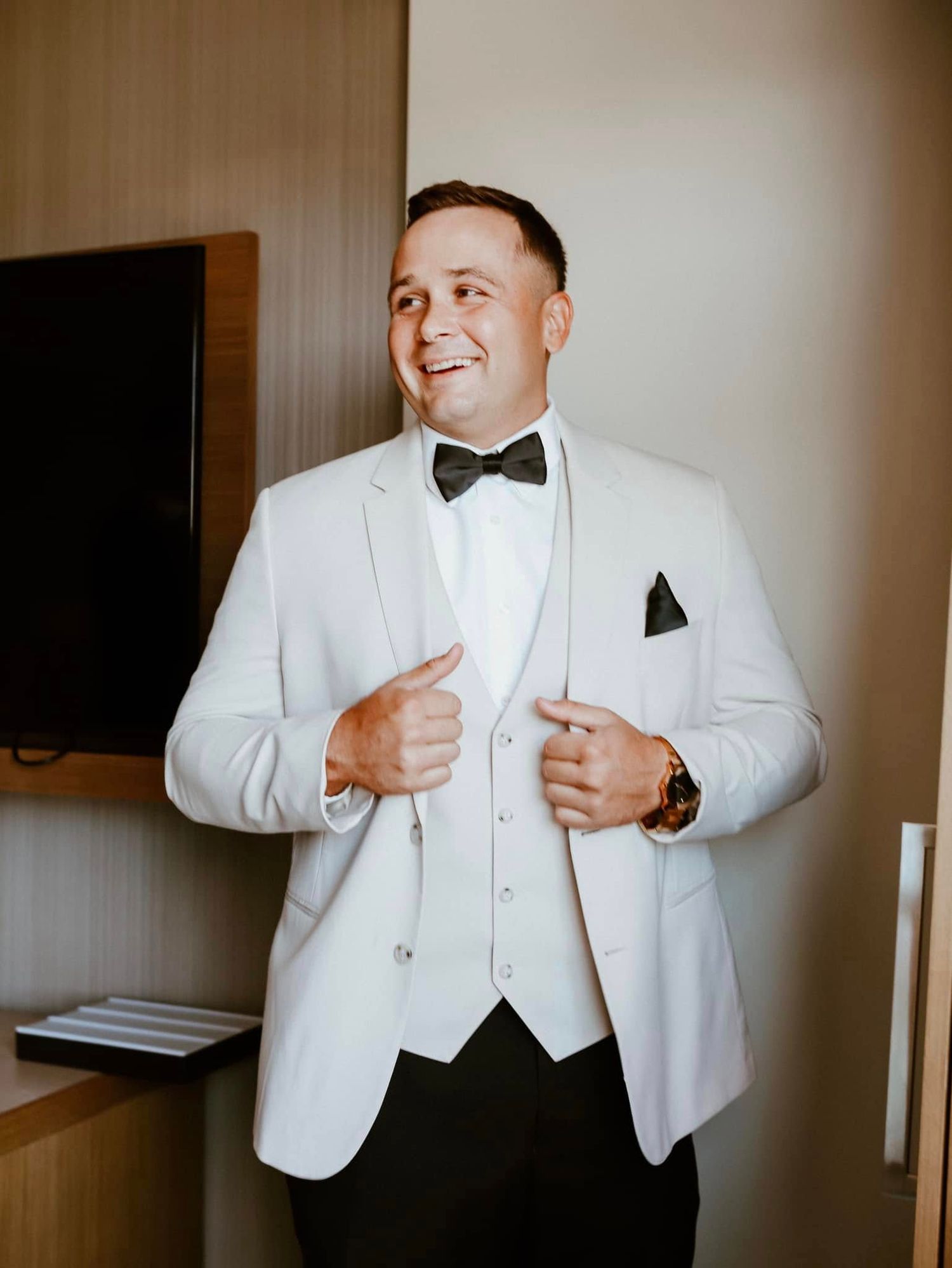 A person wearing a classic white tuxedo with black bow tie and vest adjusts their jacket while smiling.