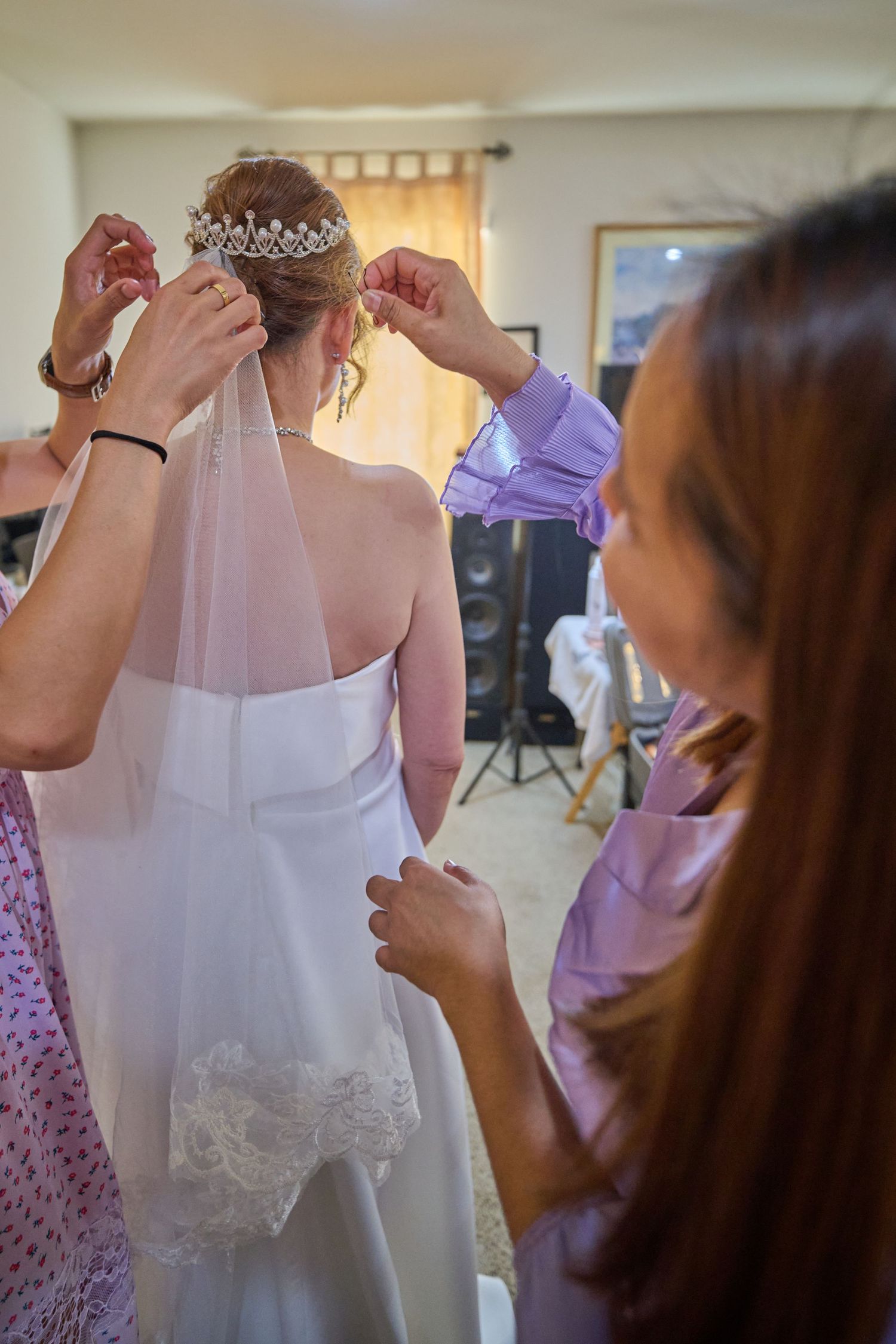 A person in a lavender shirt helps adjust a white wedding dress and veil in a bedroom setting with ceiling fan.
