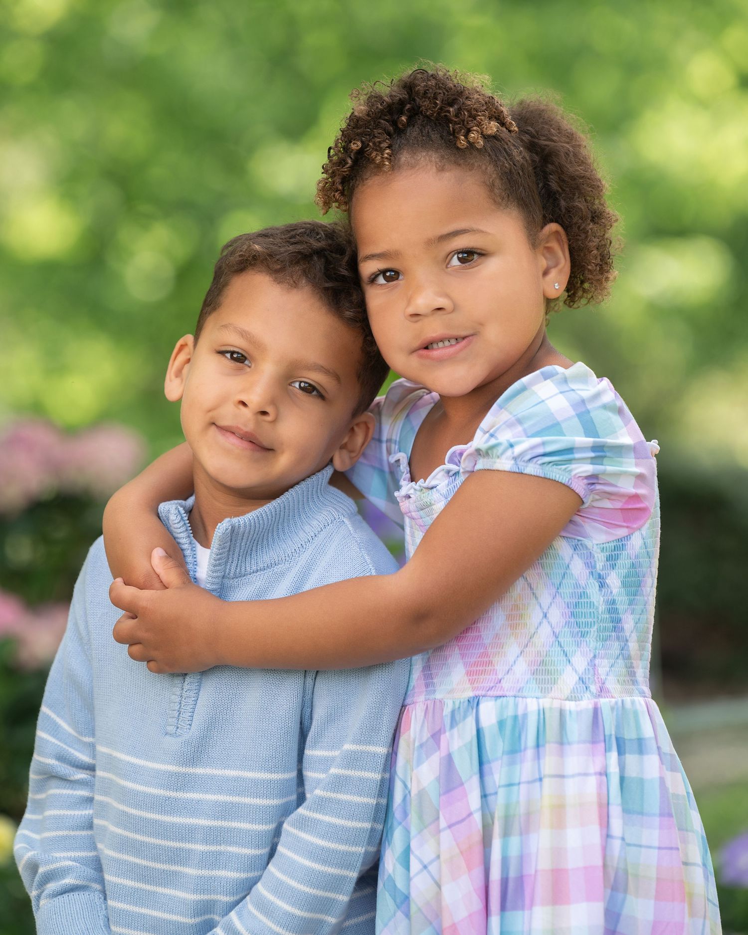 Two young children in matching striped polo shirts pose together in a garden with colorful flowers in the background.