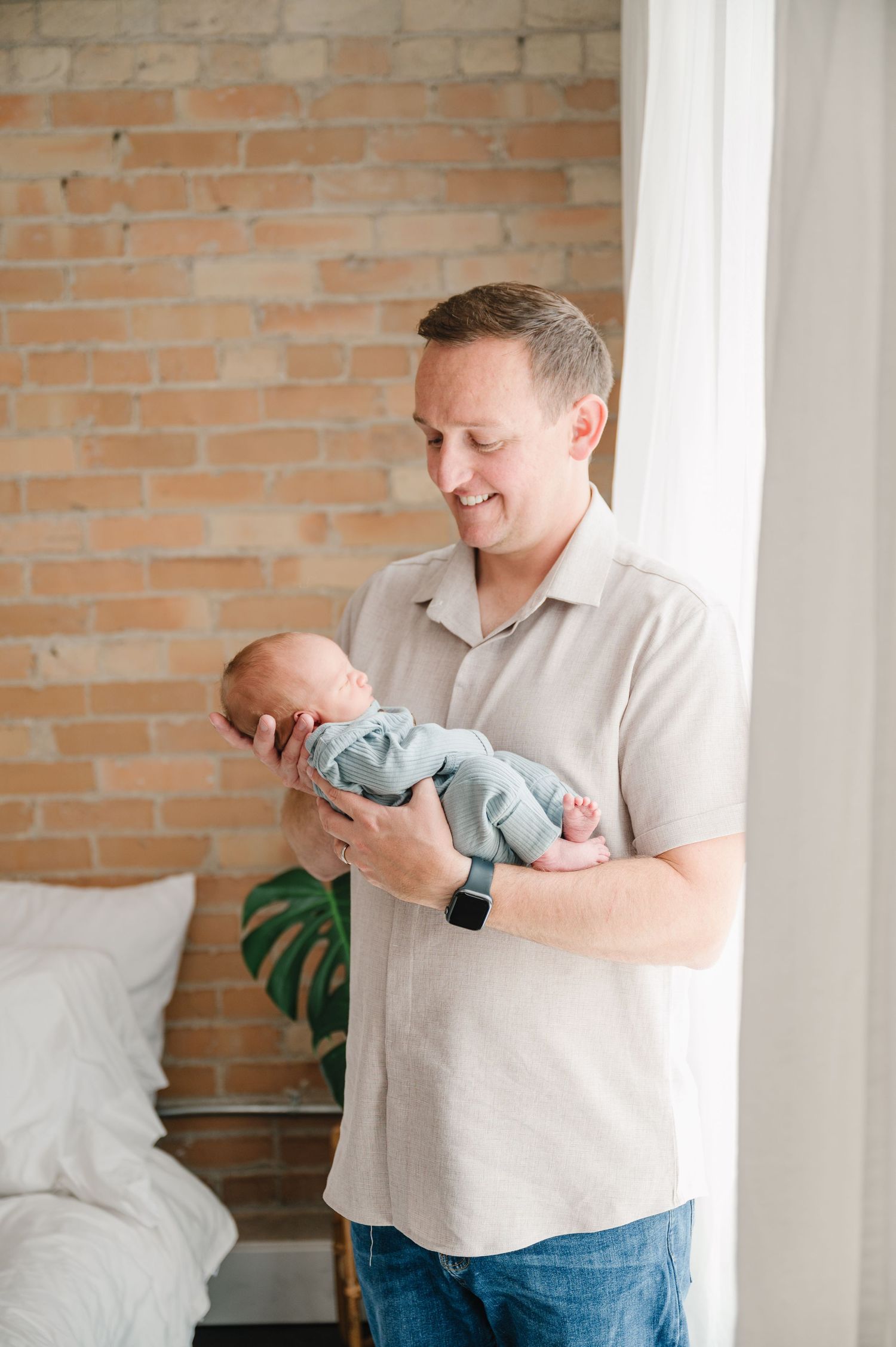 Someone holds a sleeping newborn baby wearing light blue against a brick wall backdrop in a bright room.