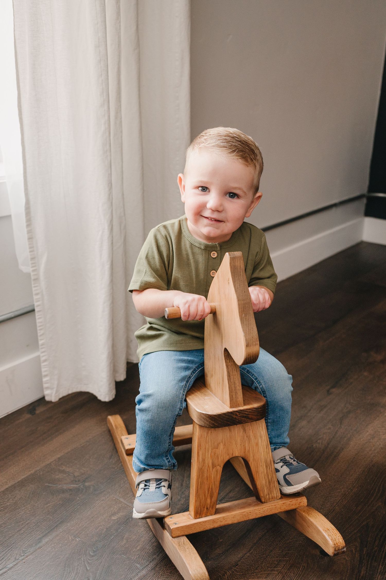 Small child wearing olive green shirt and jeans sits on wooden rocking horse toy on dark hardwood floor.