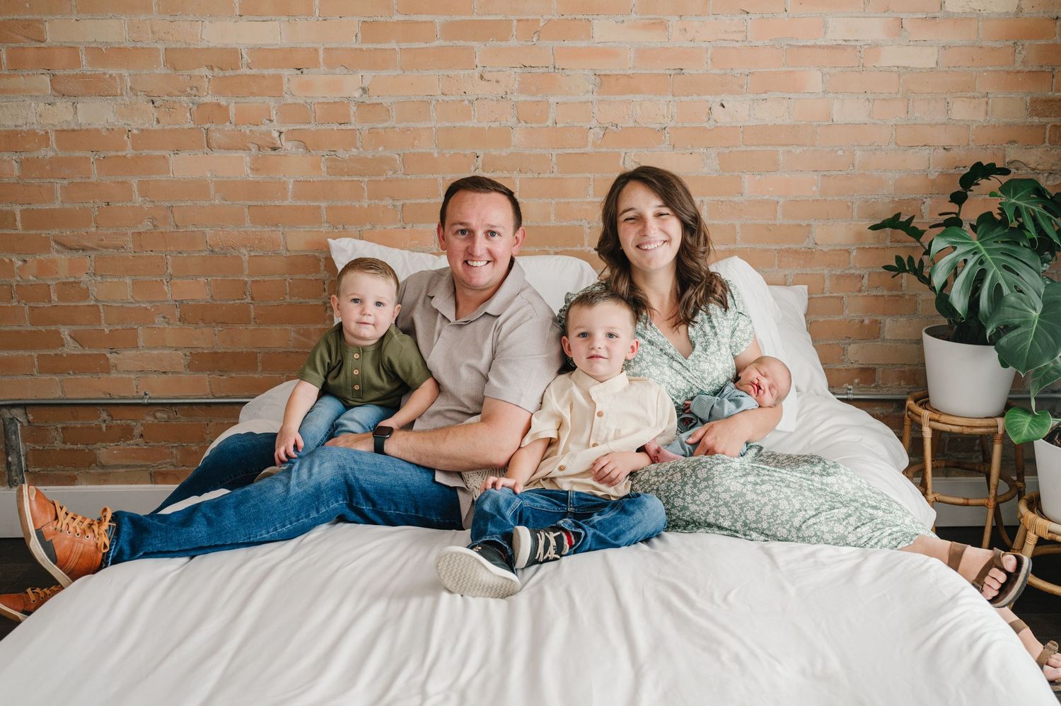Family sits together on white bedding against exposed brick wall with houseplant in corner.