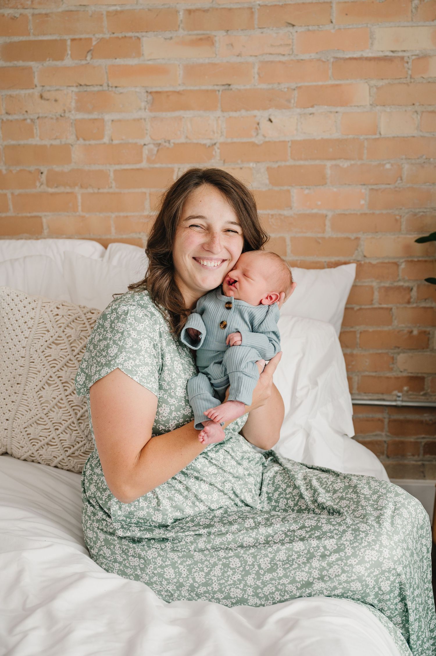 Woman in floral dress sitting on white bedding against brick wall while holding a sleeping newborn baby.