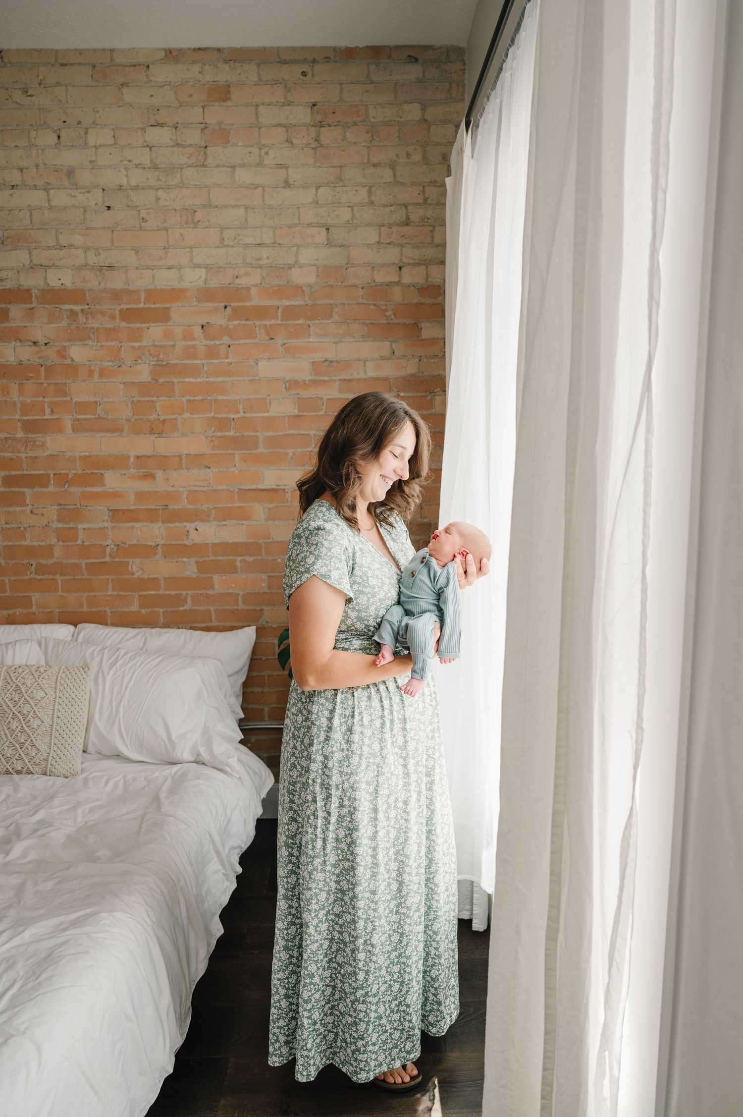 A person in a floral maxi dress stands by a window next to a white bed against an exposed brick wall.