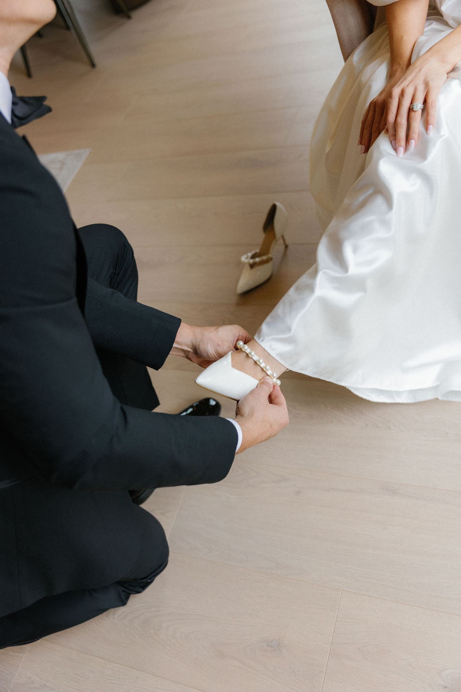 Someone in a black suit kneels on light wood flooring next to a white wedding dress and elegant high heel shoe.