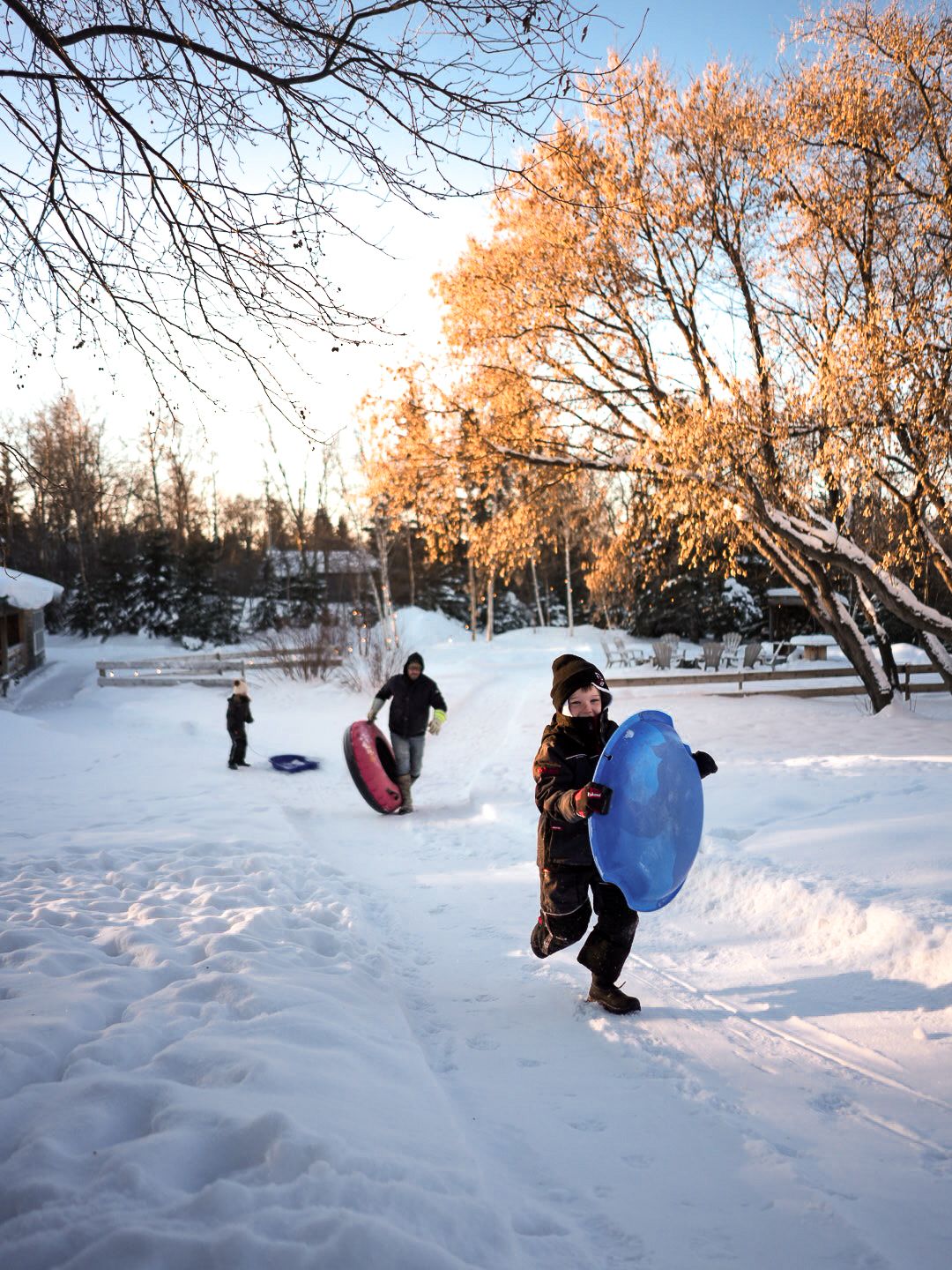 Winter scene of people playing in deep snow with bare trees at sunset.