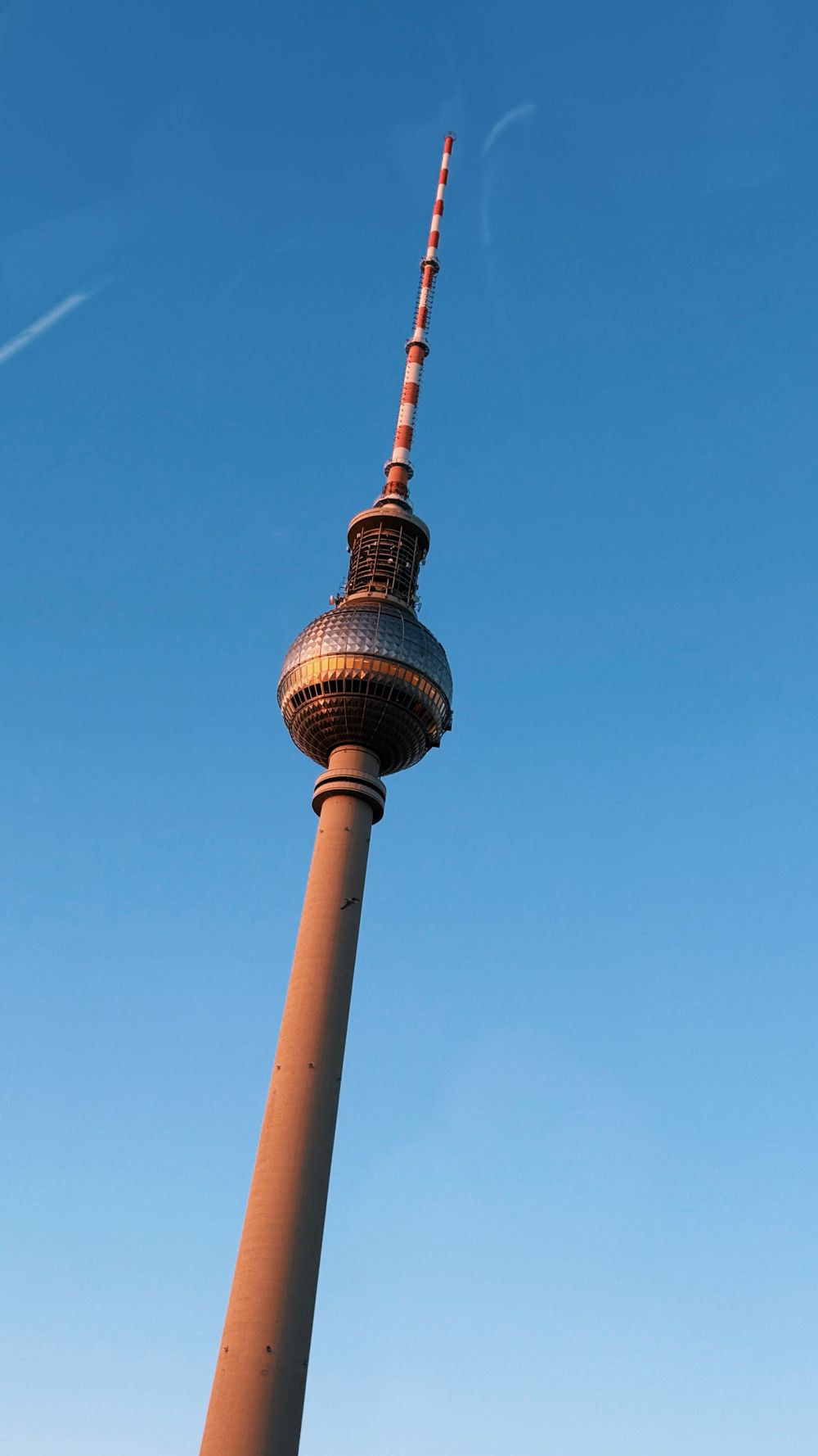 The iconic Fernsehturm television tower in Berlin rises against a bright blue sky as seen from below.