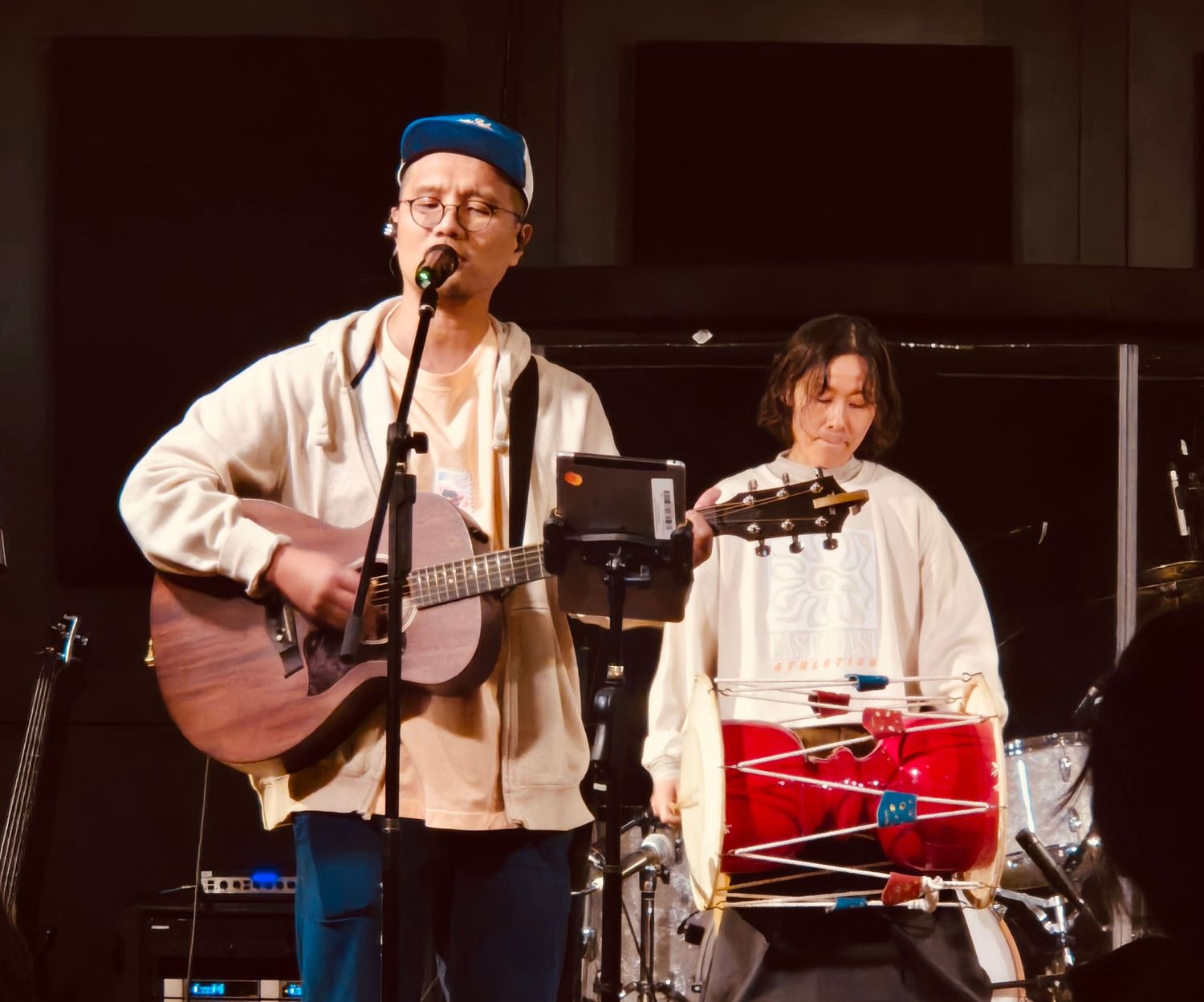 Two musicians performing live, one playing acoustic guitar and singing into a mic, another playing a red drum on stage.