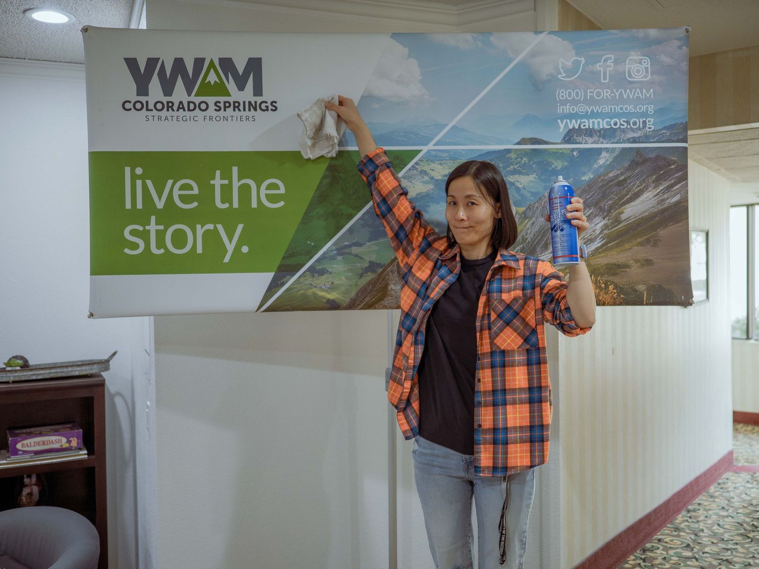 Woman smiling and pointing at a YWAM Colorado Springs 'live the story' banner in an office hallway.