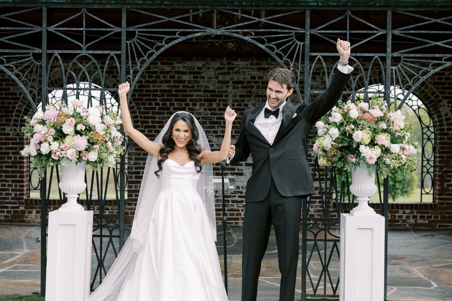Bride and groom celebrate with raised arms at elegant outdoor wedding ceremony with floral arrangements.