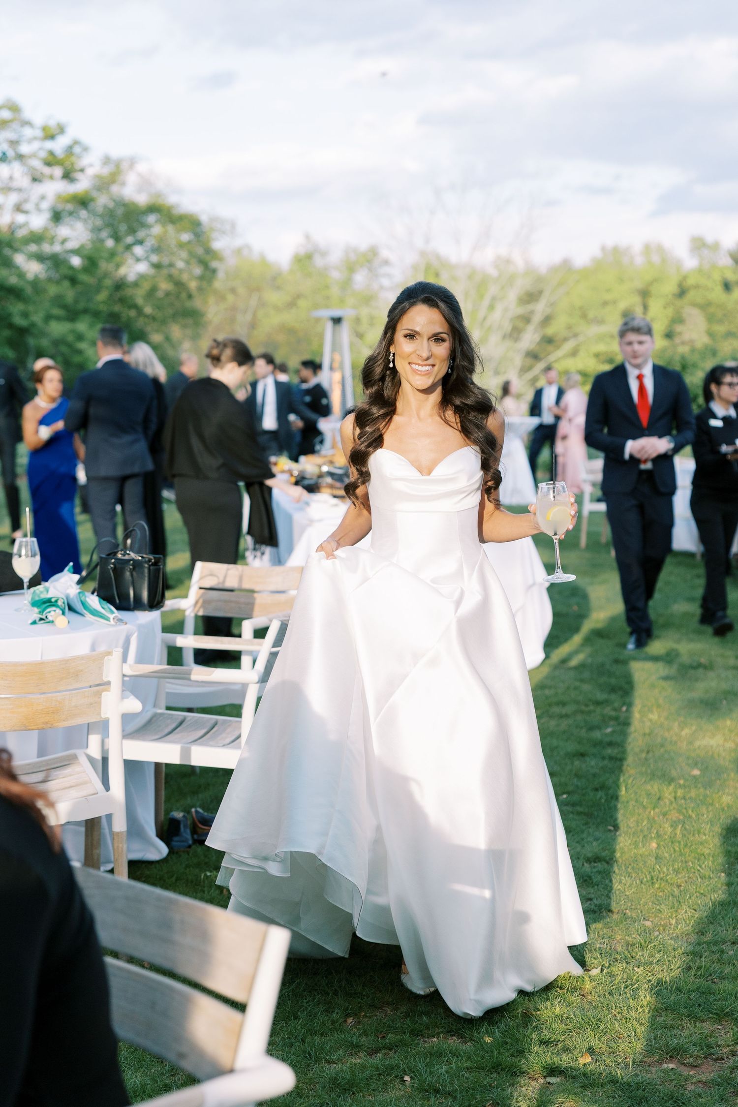 Smiling bride in a white strapless ball gown walks through an outdoor wedding reception on a green lawn.