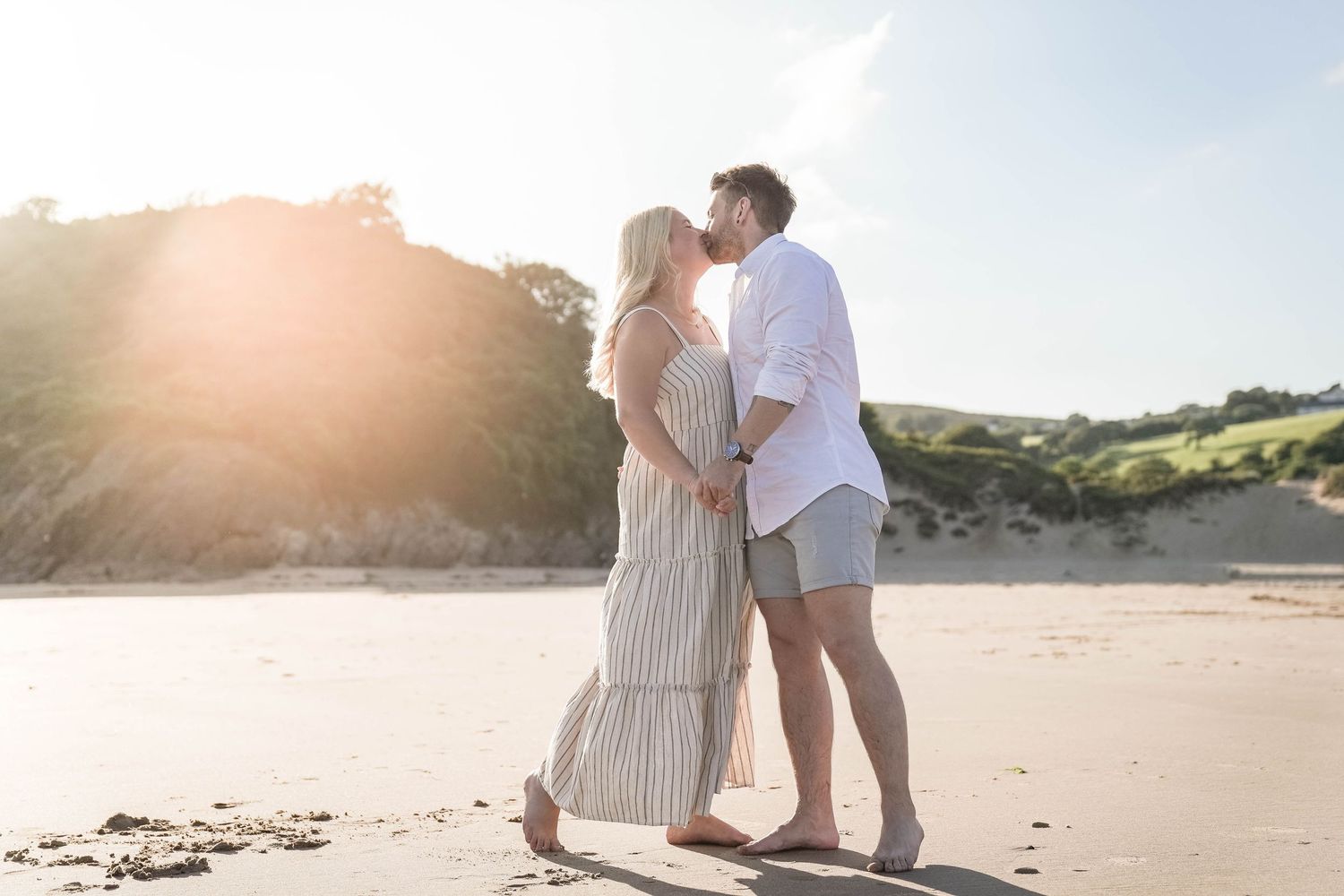 A romantic couple embraces on a sunlit beach during golden hour as sunlight streams through trees in the background.