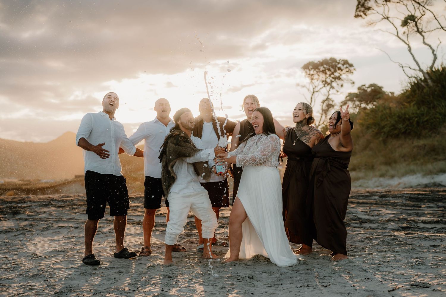 Group of friends celebrating on a beach at sunset, with two people in white dresses popping champagne.