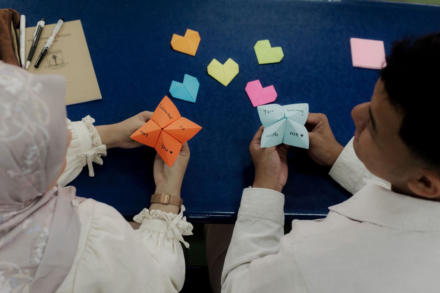 Two students making colorful origami paper fortune tellers with heart shapes on a blue bulletin board.