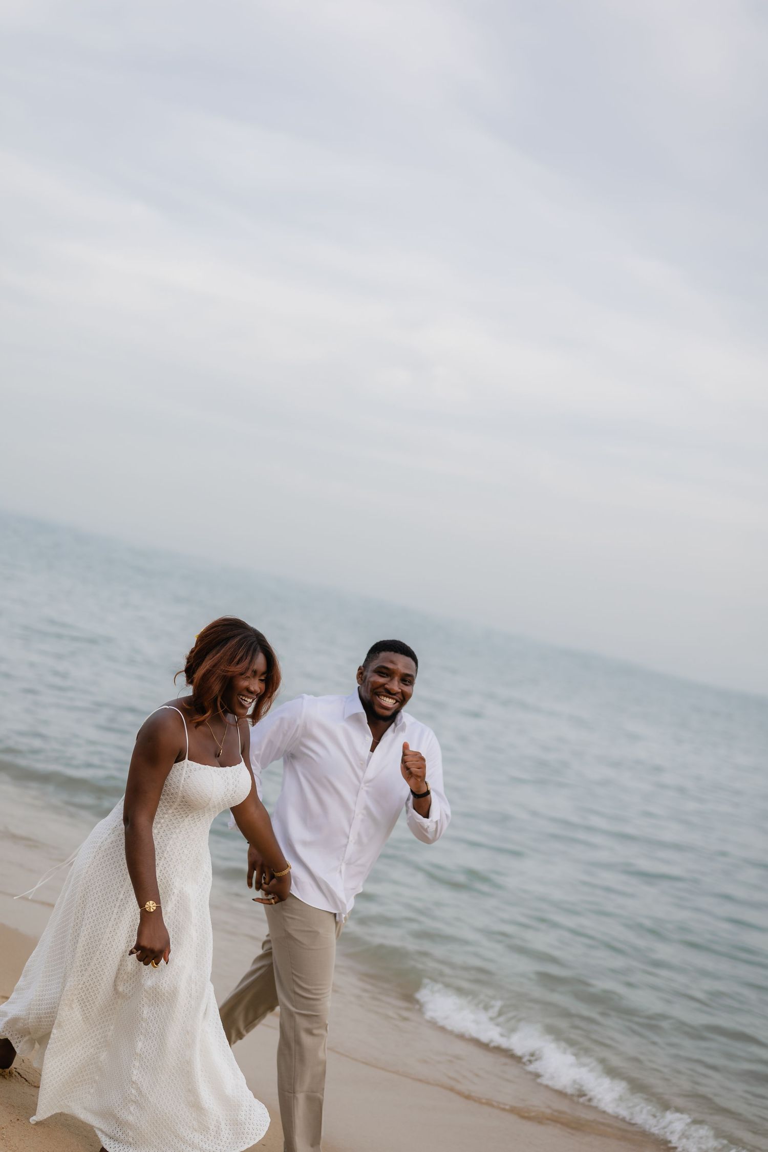 Happy couple in white attire walking hand in hand on a sandy beach with calm ocean waves in the background.