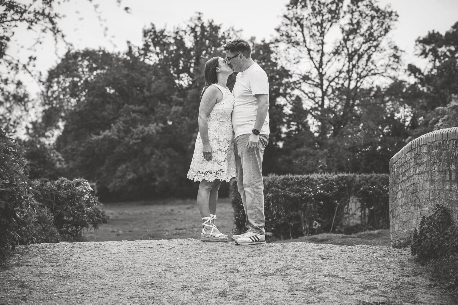 Black and white photo of a couple sharing a romantic moment on a stone bridge surrounded by lush trees in a park.