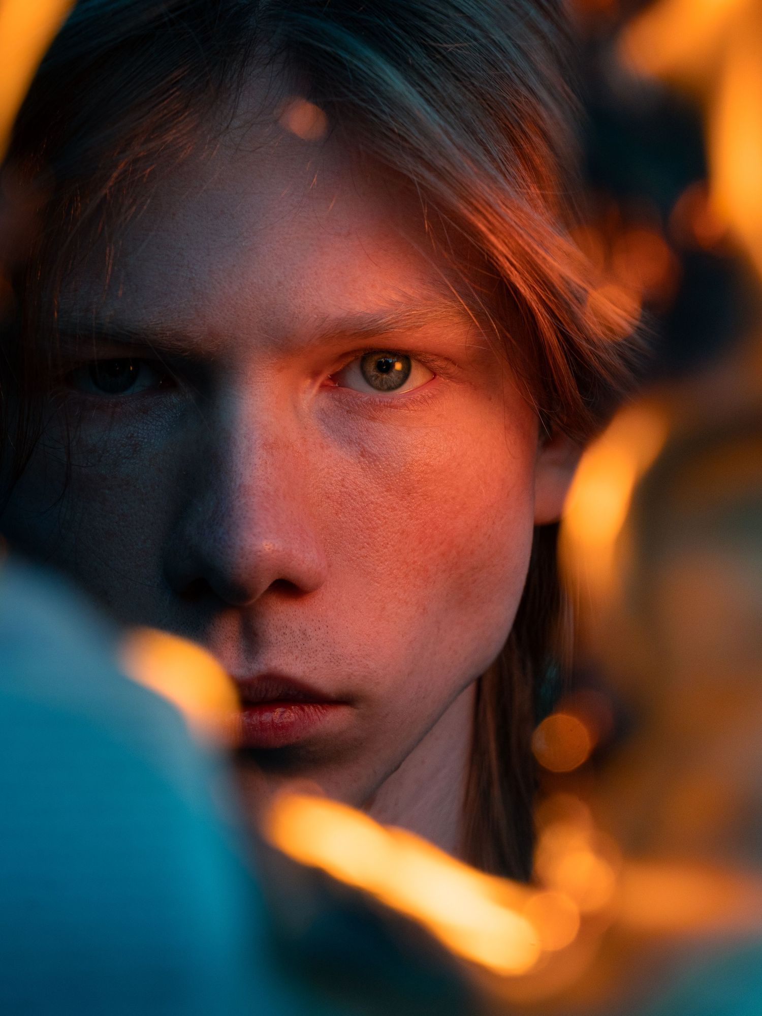 Close-up portrait of a young person with long hair, illuminated by warm golden bokeh lights in a moody setting.
