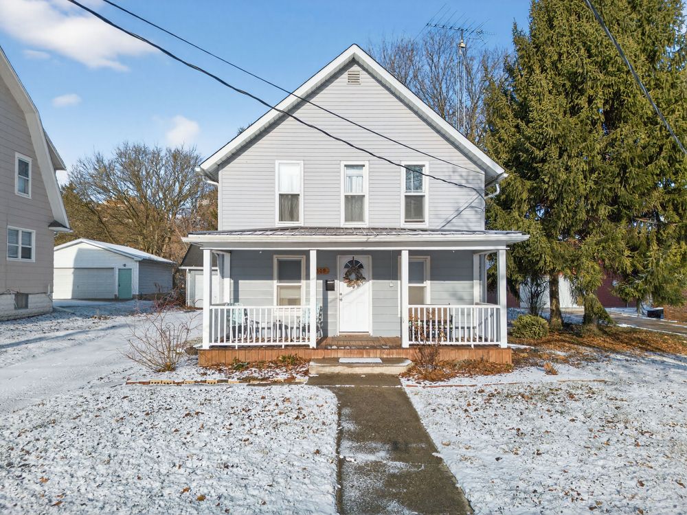 Gray two-story farmhouse with white porch railing, snow-covered yard, and large evergreen tree in winter setting.