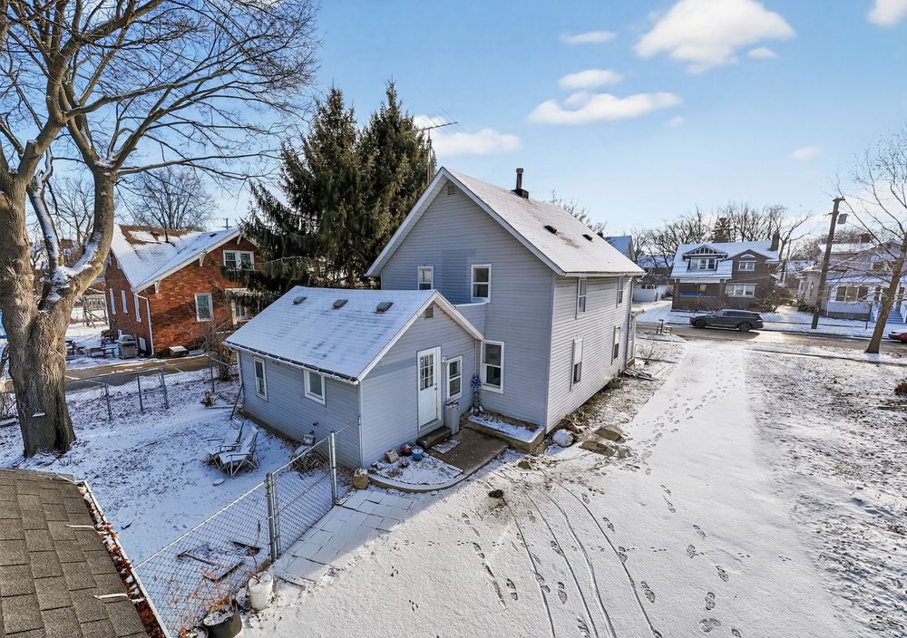 Aerial view of a light blue two-story home surrounded by snow-covered ground and evergreen trees in winter.