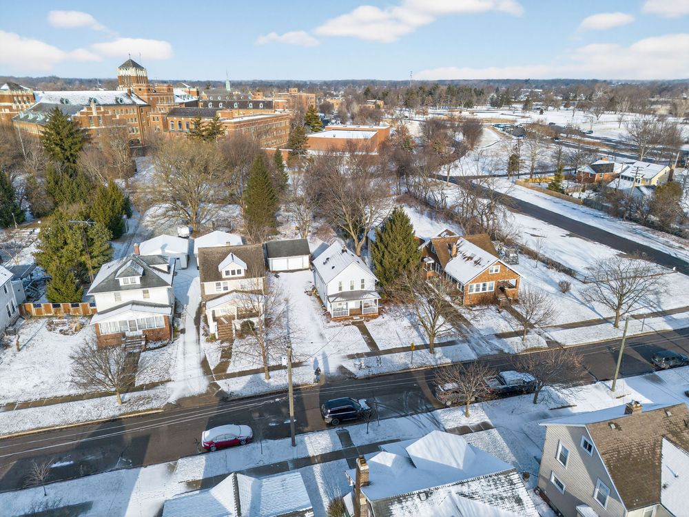 Snowy aerial view of a residential neighborhood with bare trees, homes, and a large brick building in the background.