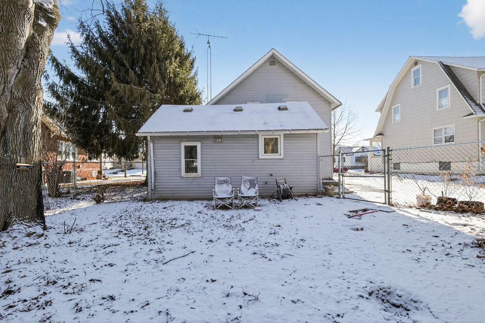 Snow-covered backyard of a small gray house with two chairs, a fence, and bare trees in a residential neighborhood.