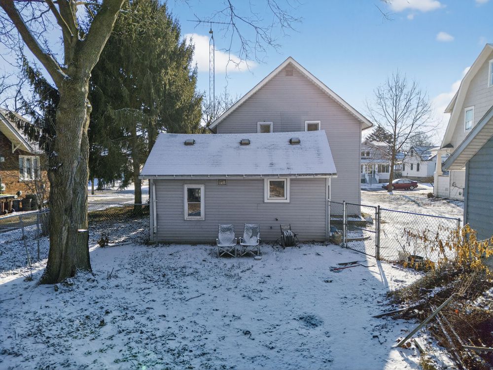 Snow-covered backyard of a gray two-story home with outdoor chairs, large trees, and neighboring houses visible in winter.