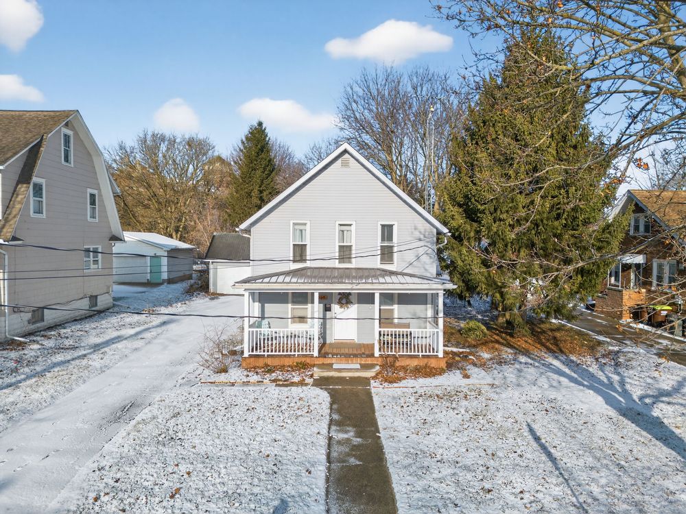 White two-story farmhouse with covered porch surrounded by snow-covered yard and large evergreen trees.