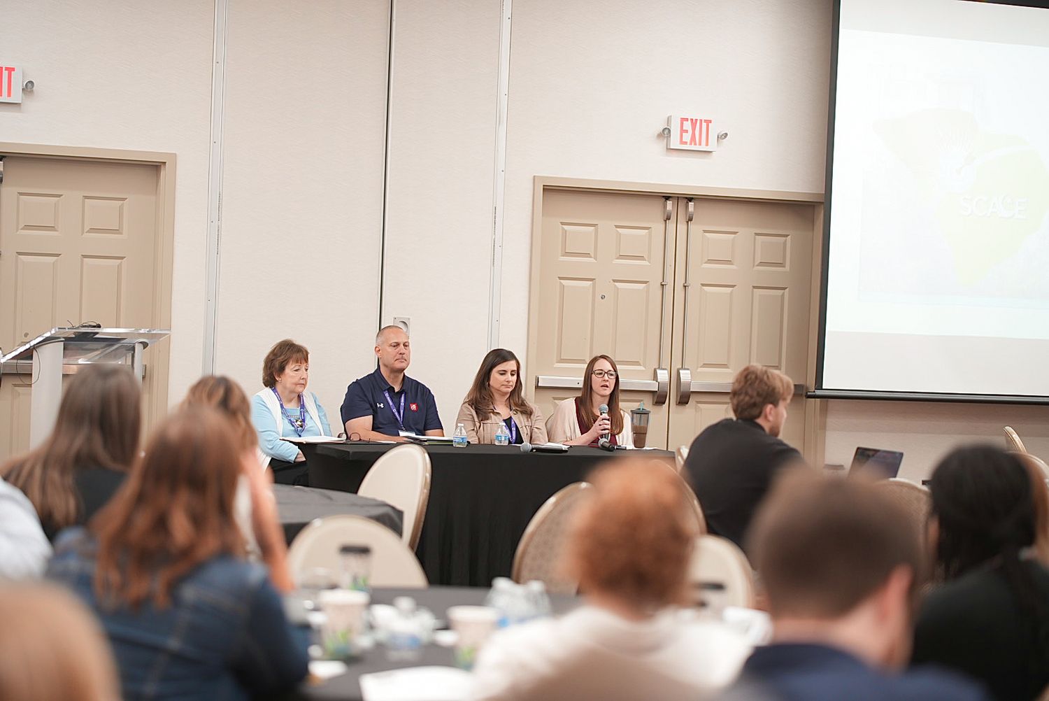 Four panelists sit at a draped table facing an audience seated at round tables during a conference session in a meeting room.