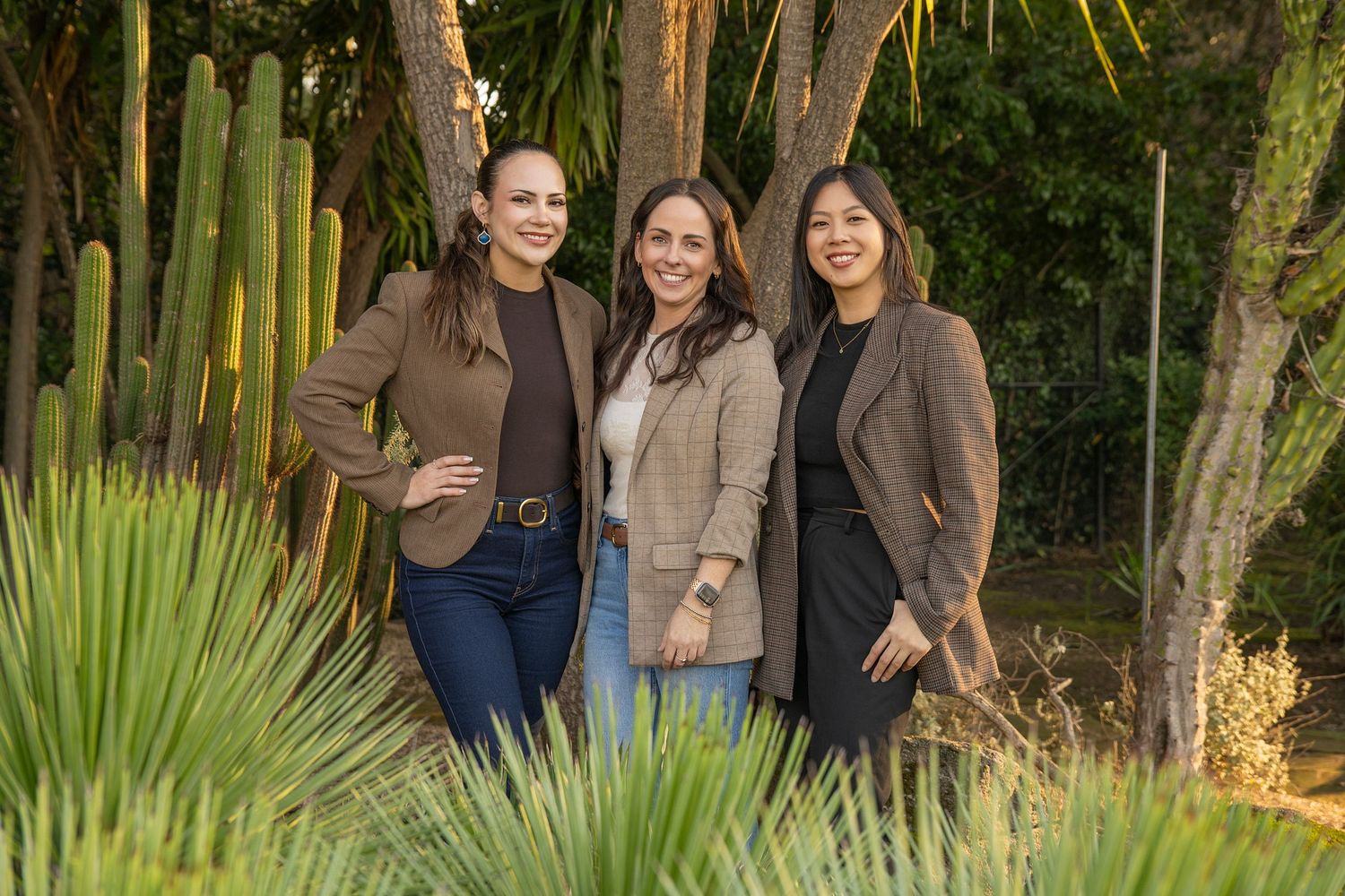 Three women in brown blazers smiling outdoors among tropical plants and tall cacti in a lush garden setting.