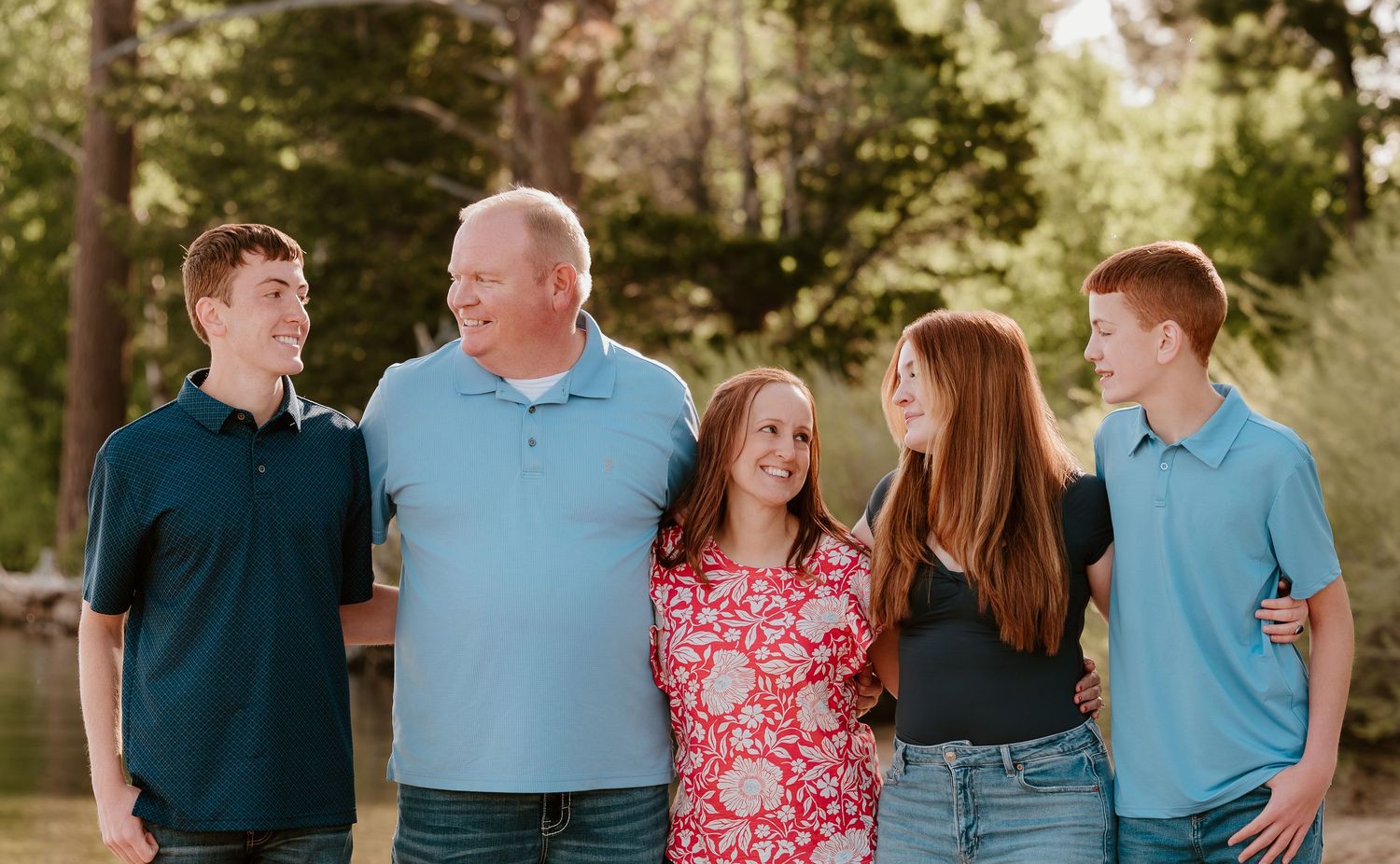 Happy family of five smiling outdoors in a park setting with trees in the background.