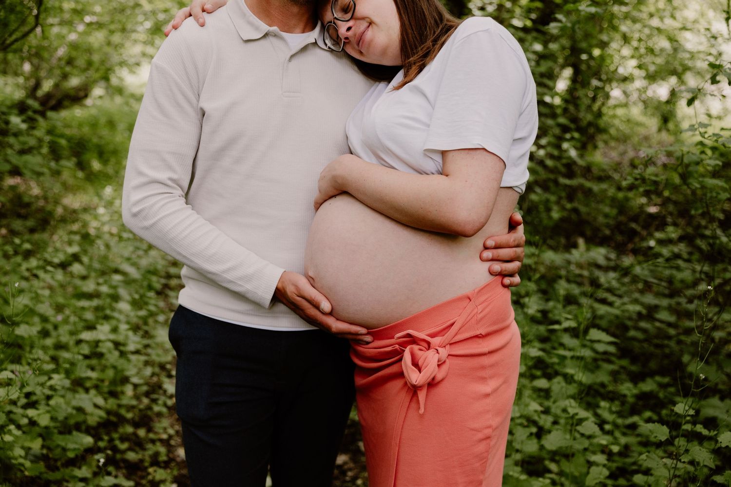 Couple tenderly embracing outdoors, showing pregnant belly surrounded by lush green foliage.