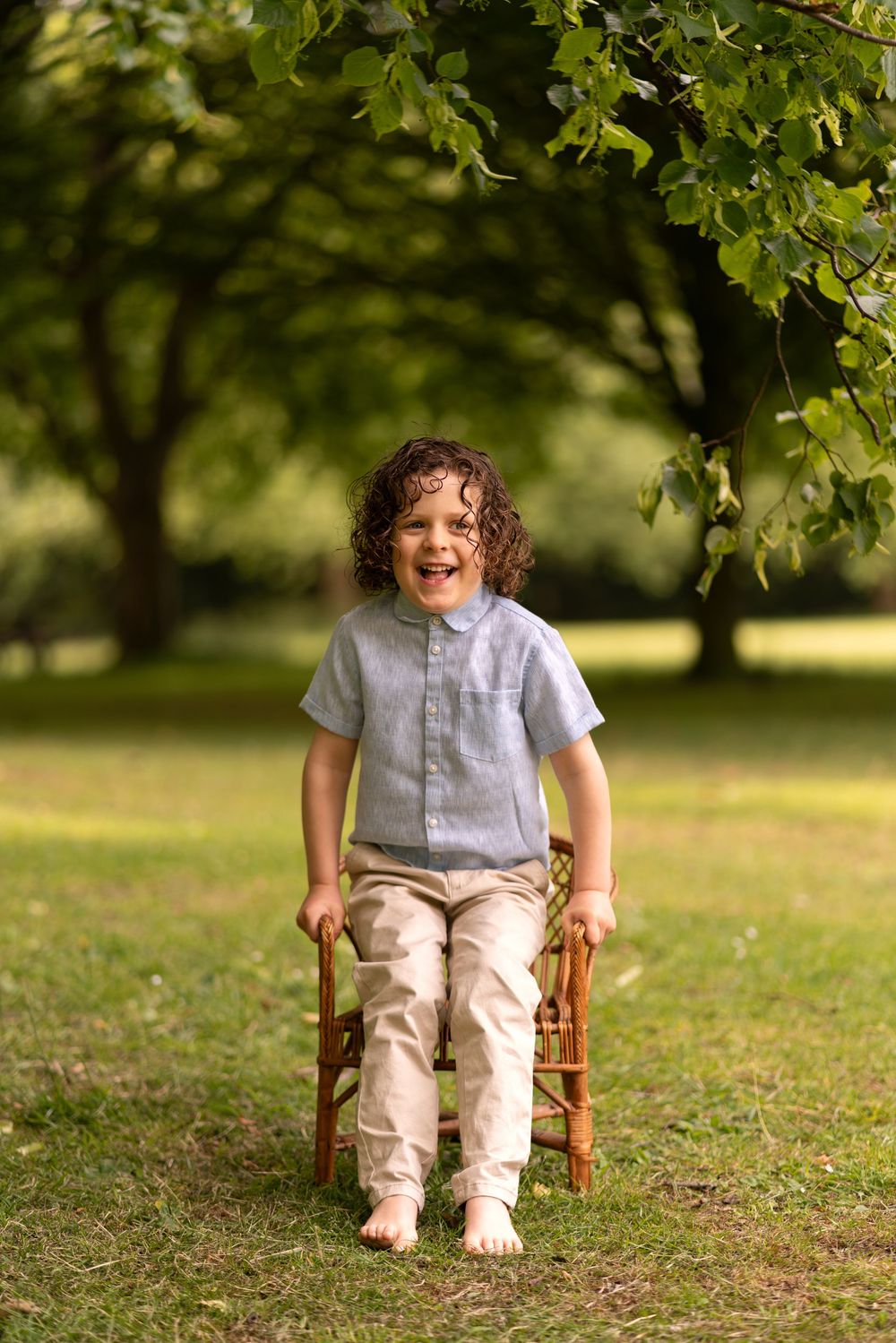 A joyful curly-haired boy sits on a wooden chair barefoot in a lush green park, wearing a grey shirt and beige pants.